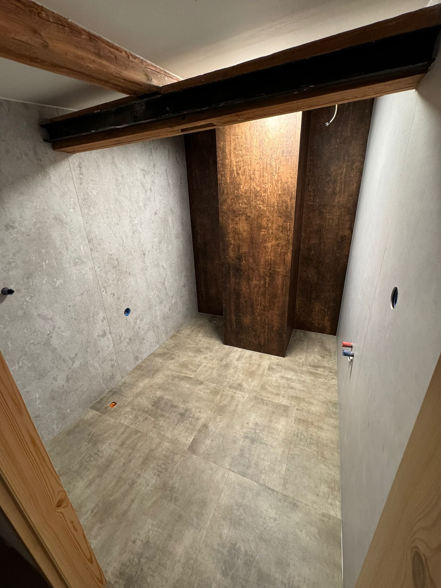 Wnętrze łazienki w trakcie remontu z szarymi płytkami na ścianach i podłodze, drewnianymi belkami stropowymi i pionowymi panelami w odcieniach brązu, widoczne otwory na instalacje.