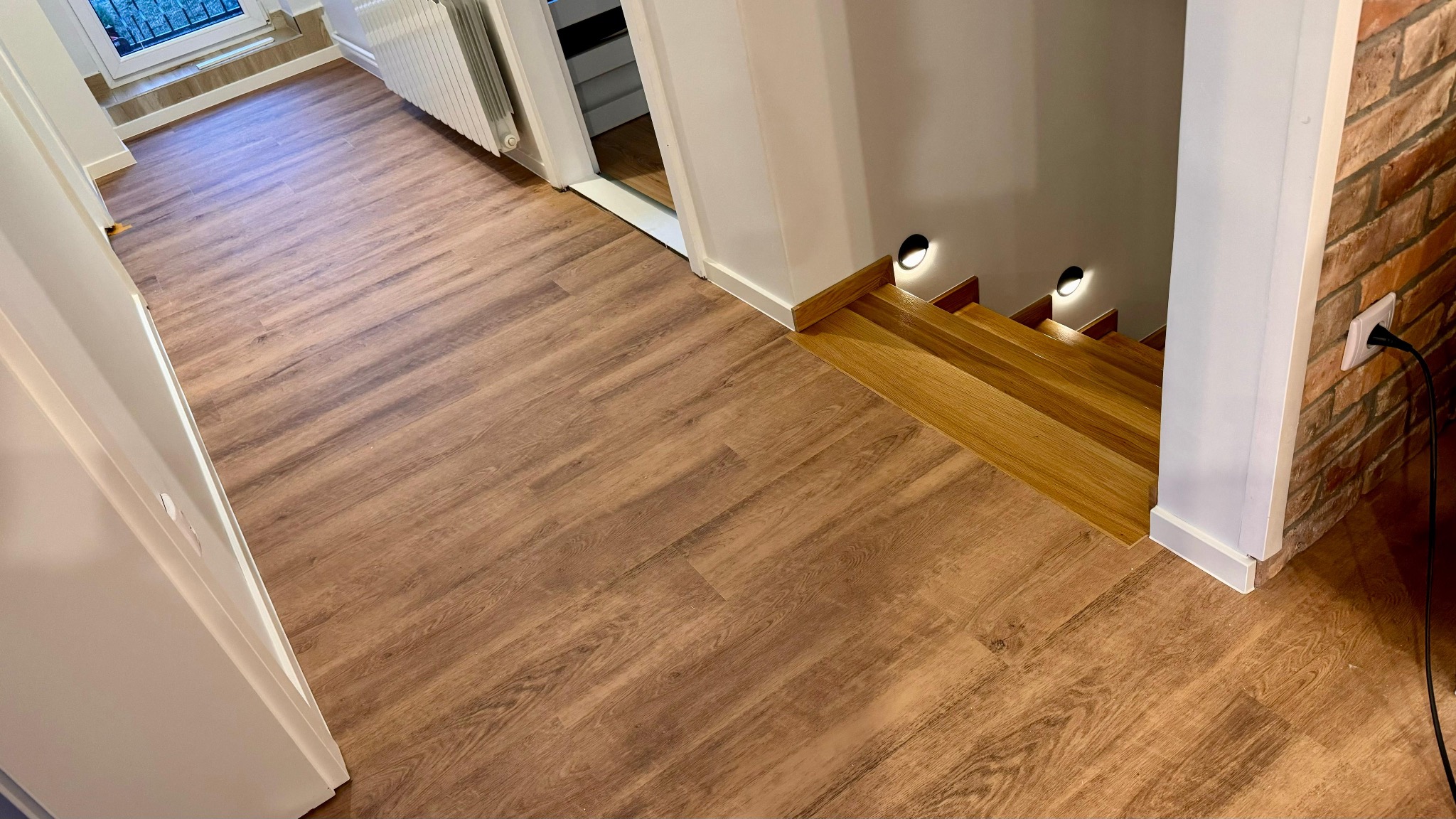 Ukończona podłoga z paneli winylowych o wzorze drewna, prowadząca do drewnianych schodów z oświetleniem punktowym i ceglanej ściany z gniazdkiem elektrycznym.