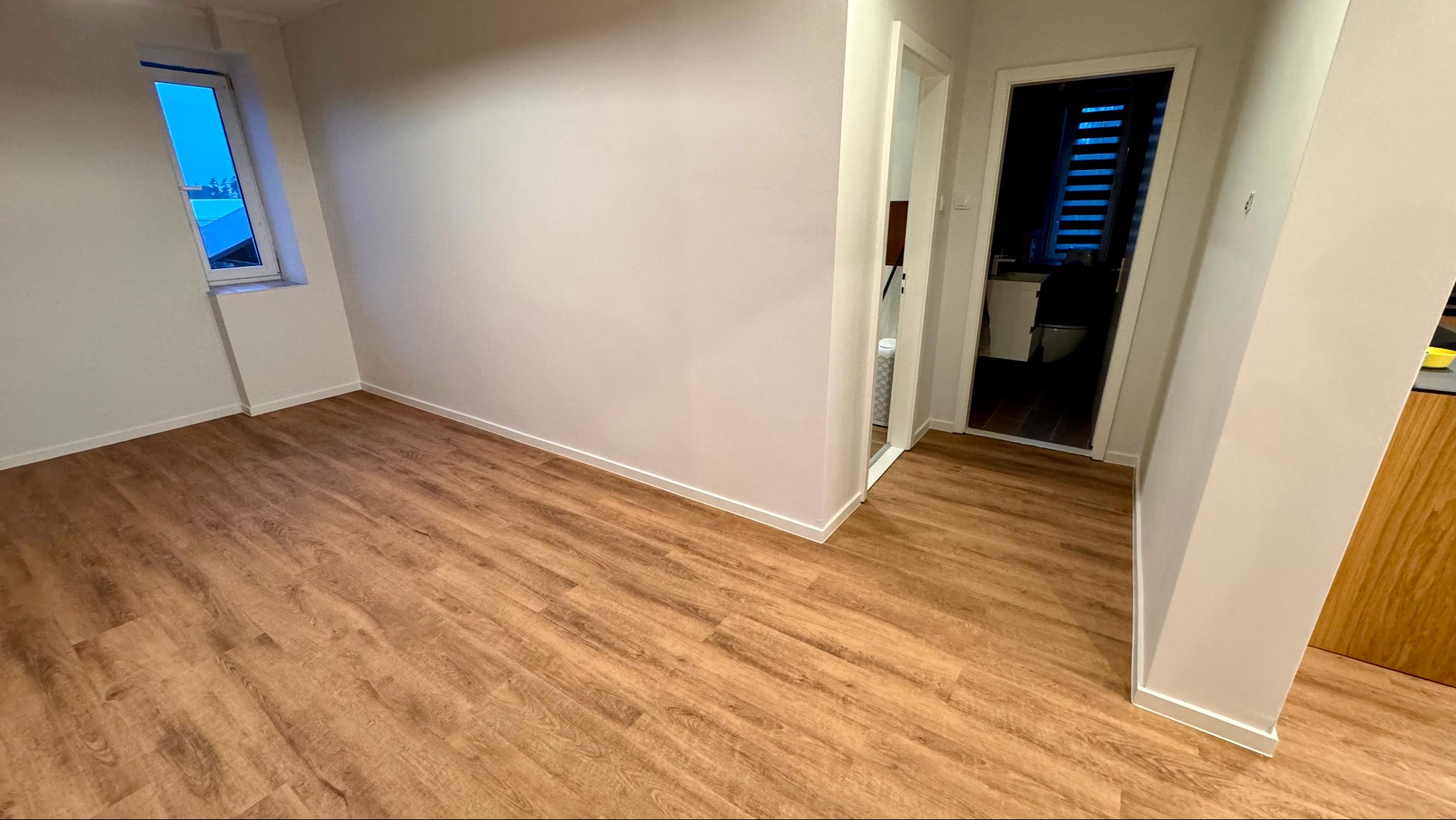 Jasna, pusta przestrzeń z nową podłogą imitującą drewno ułożoną w jodełkę, widok na okno i wejście do łazienki.