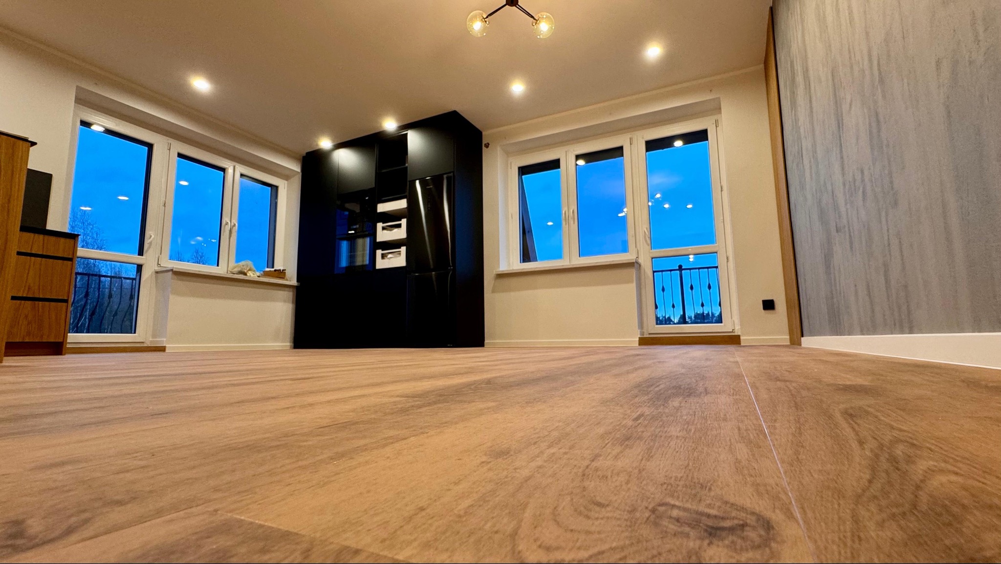 Jasne panele podłogowe w nowym mieszkaniu z widokiem na nocne niebo za oknem balkonowym i czarną zabudową kuchenną w tle.