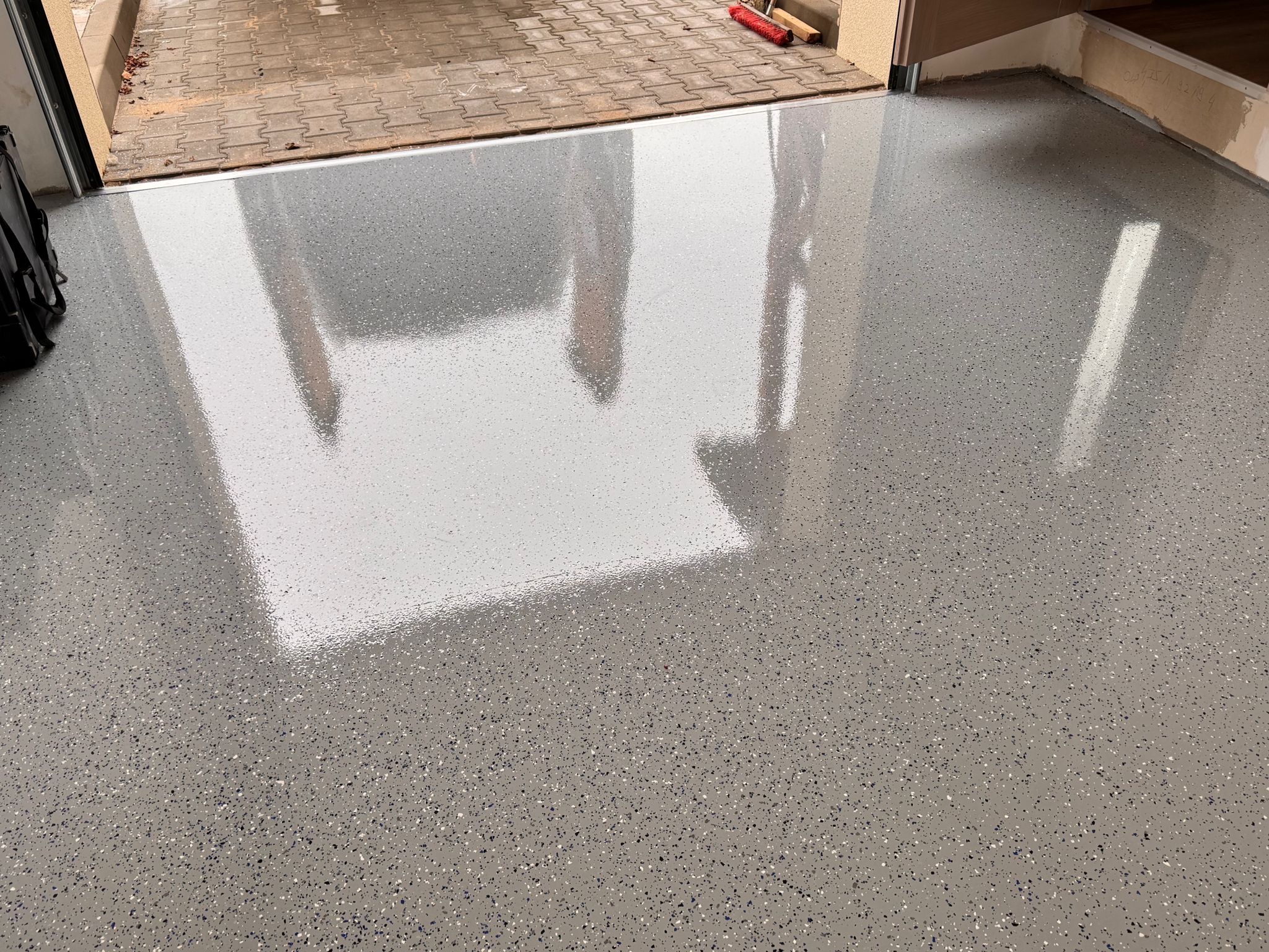Błyszcząca, szara posadzka z drobnymi, kolorowymi wtrąceniami, odbijająca światło w garażu z otwartą bramą na zewnątrz.