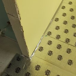 Andro-Bud - Przygotowanie do montażu paneli podłogowych z folią izolacyjną z nadrukowanymi liniami cięcia na nierównej podłodze w rogu pomieszczenia, widoczna ściana z surowym wykończeniem.