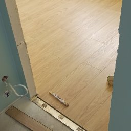 Andro-Bud - Układanie paneli podłogowych w pokoju z żółtą ścianą, widoczny fragment ściany z gniazdkiem, listwa przypodłogowa w trakcie montażu, miarka stolarska leży na podłodze.