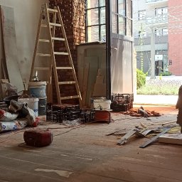 Andro-Bud - Wnętrze w trakcie prac we Wrocławiu: fragment ściany z cegły z krzyżykami dystansowymi, drewniana drabina, worki z zaprawą, otwarte okno z widokiem na budynek mieszkalny.