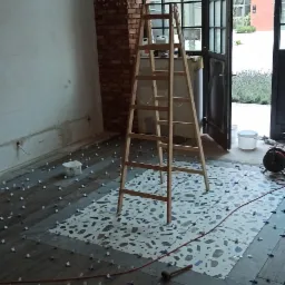 Układanie mozaiki podłogowej z użyciem krzyżyków dystansowych; widoczna drewniana drabina, fragment ściany z cegły i otwarte szklane drzwi.