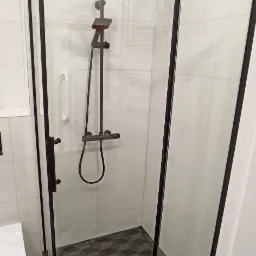 Nowoczesna kabina prysznicowa z czarną ramą i deszczownicą, białe płytki ścienne, podłoga z geometrycznym wzorem 3D, uchwyt dla niepełnosprawnych.