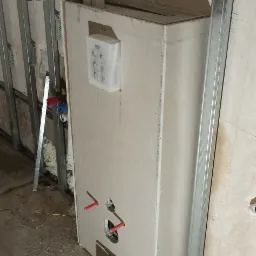 Obudowa stelaża podtynkowego WC z płyt kartonowo-gipsowych w trakcie montażu, widoczne profile metalowe, wystające rury instalacji wodnej oraz karton z płytkami Grohe. Na obudowie widoczna...