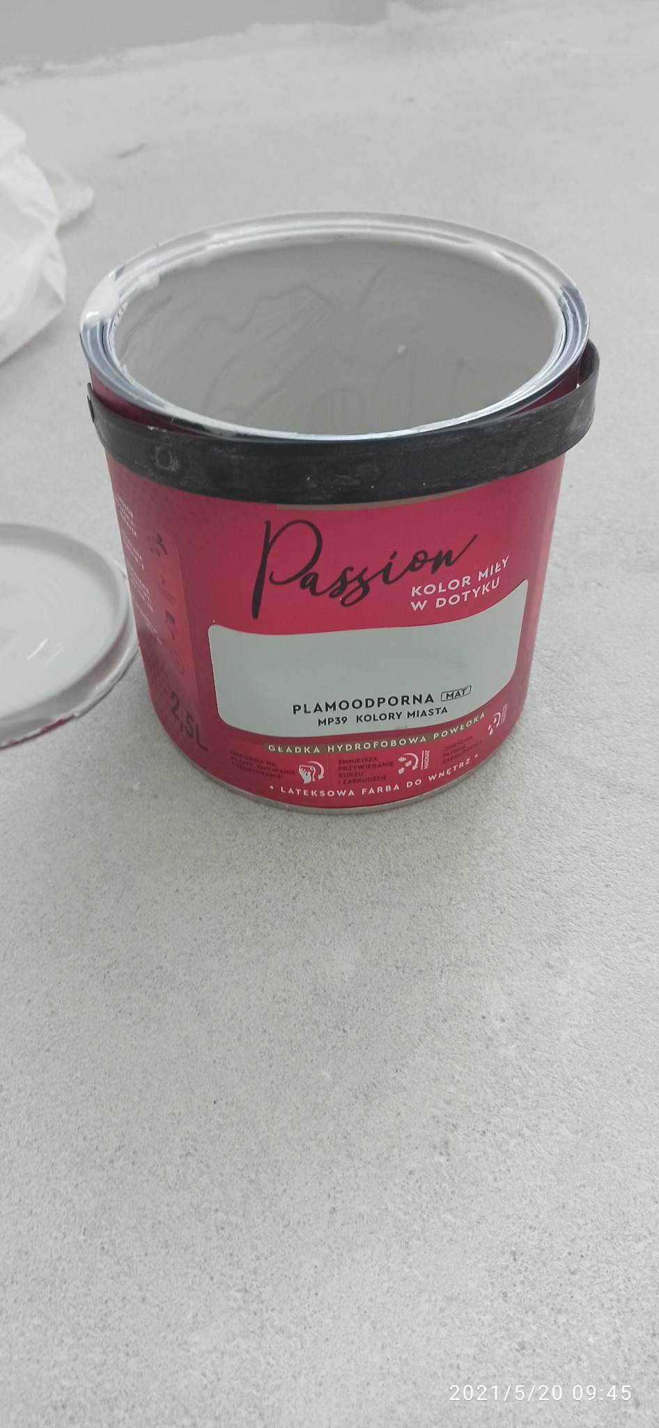 Otwarta puszka farby lateksowej 'Passion' w kolorze jasnoszarym, z widocznym wieczkiem obok, na tle szarej, surowej powierzchni.