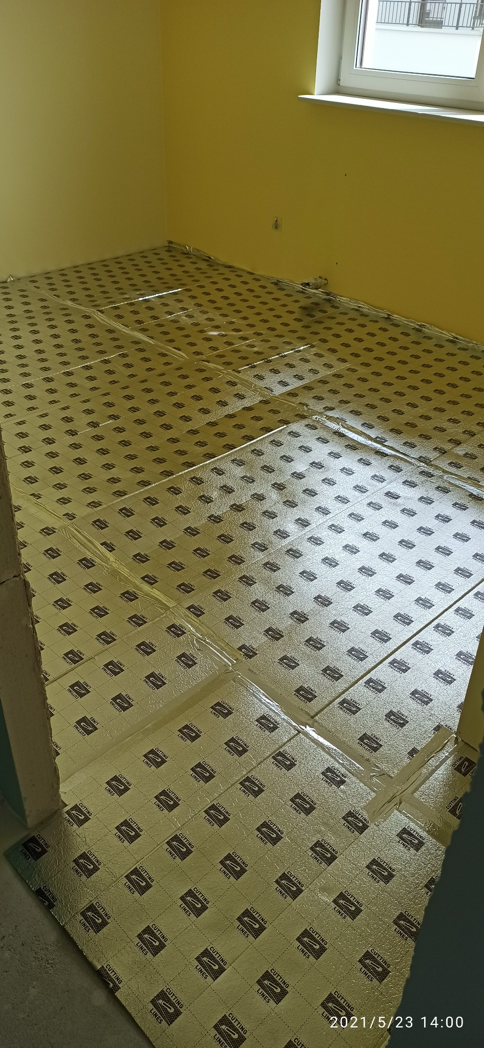 Przygotowanie podłoża pod posadzkę w pokoju z żółtymi ścianami, wyłożone maty izolacyjne z napisem 'CUTTING LINES', widoczne okno i fragment ściany z niedokończonym wykończeniem.