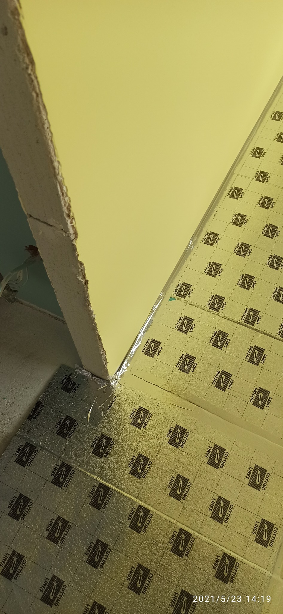 Przygotowanie do montażu paneli podłogowych z folią izolacyjną z nadrukowanymi liniami cięcia na nierównej podłodze w rogu pomieszczenia, widoczna ściana z surowym wykończeniem.