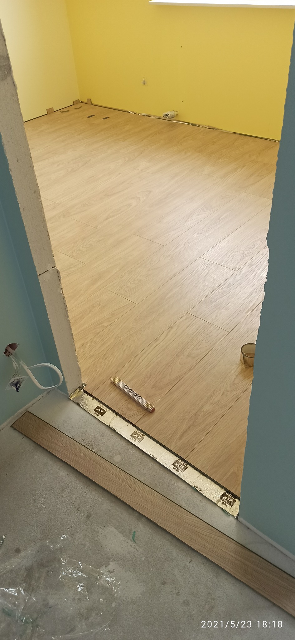 Układanie paneli podłogowych w pokoju z żółtą ścianą, widoczny fragment ściany z gniazdkiem, listwa przypodłogowa w trakcie montażu, miarka stolarska leży na podłodze.