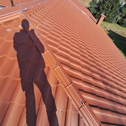 JG ELEKTRO SPÓŁKA Z OGRANICZONĄ ODPOWIEDZIALNOŚCIĄ - Czerwona dachówka z zamontowaną instalacją odgromową, widok z góry z cieniem osoby, w tle zielony krajobraz.