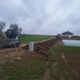 JG ELEKTRO SPÓŁKA Z OGRANICZONĄ ODPOWIEDZIALNOŚCIĄ - Wykopany rów w ziemi pod instalacje, z widocznym minikoparką na przyczepie i szarą szafką elektryczną. W tle dom i szklarnie.