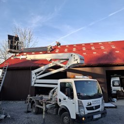 JG ELEKTRO SPÓŁKA Z OGRANICZONĄ ODPOWIEDZIALNOŚCIĄ - Montaż paneli słonecznych na czerwonym dachu budynku gospodarczego z użyciem podnośnika koszowego. Widoczny samochód dostawczy firmy MetalStacja.