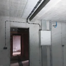Surowy beton i instalacje elektryczne: kable w peszlach na suficie, puszka rozdzielcza na ścianie, widok z wnętrza budynku w stanie deweloperskim.