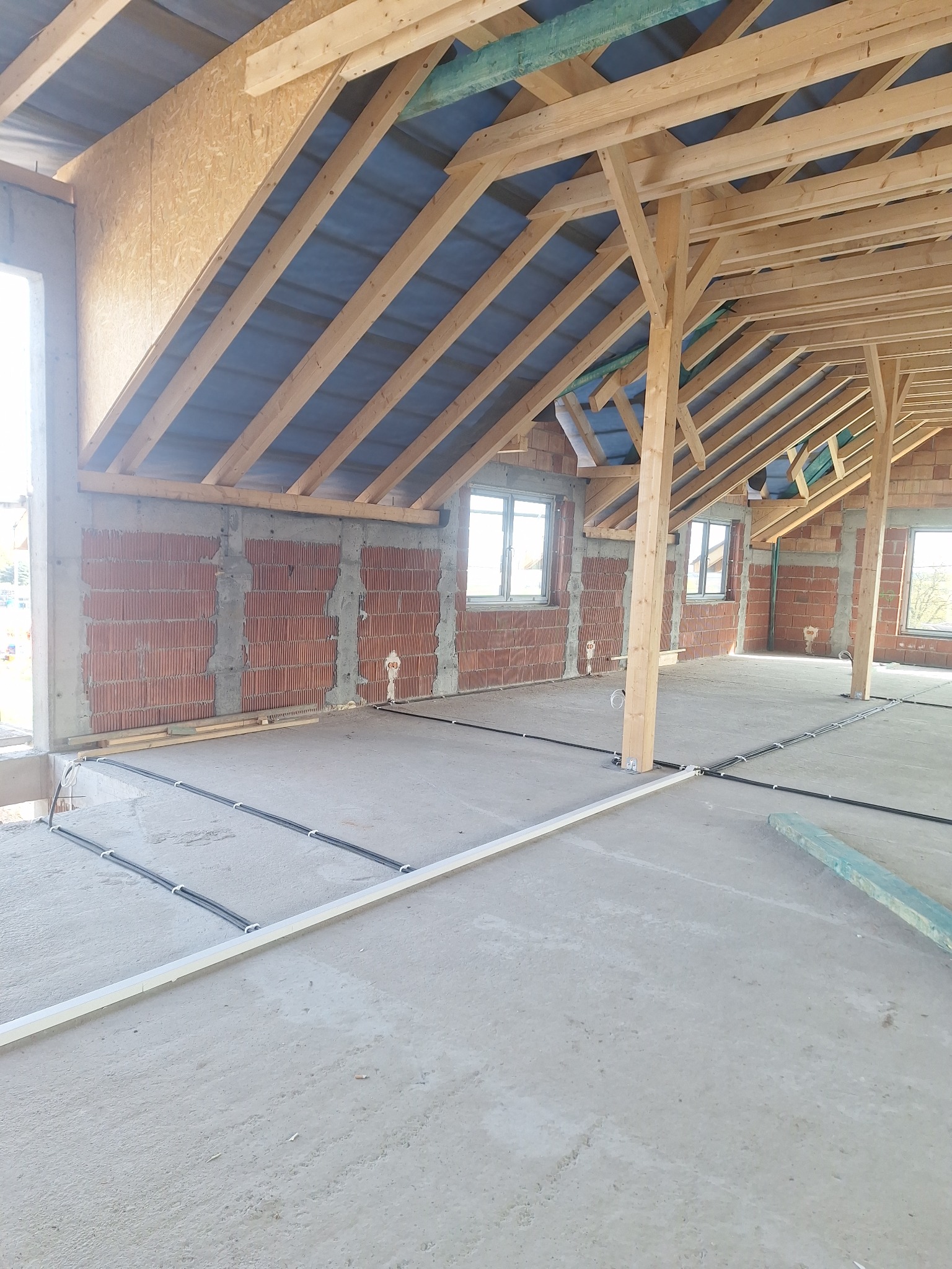 Wnętrze budynku w trakcie budowy: widoczne mury z cegły, drewniana konstrukcja dachu i podłoga z rozprowadzonymi przewodami elektrycznymi w peszlach.