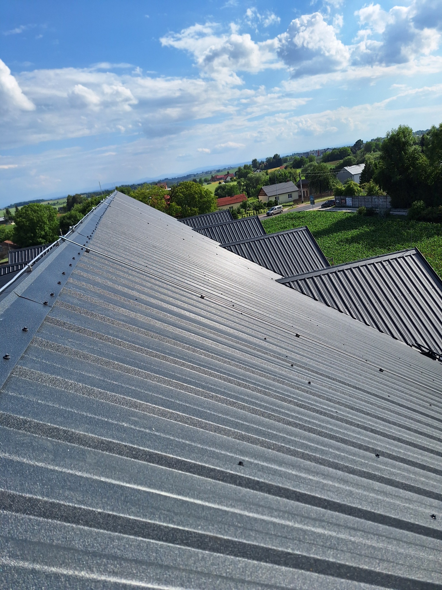 Szara blacha trapezowa na dachu z widokiem na okolicę Koniuszy. Widoczne mocowania i system odgromowy. Dach pokryty blachą w słoneczny dzień.