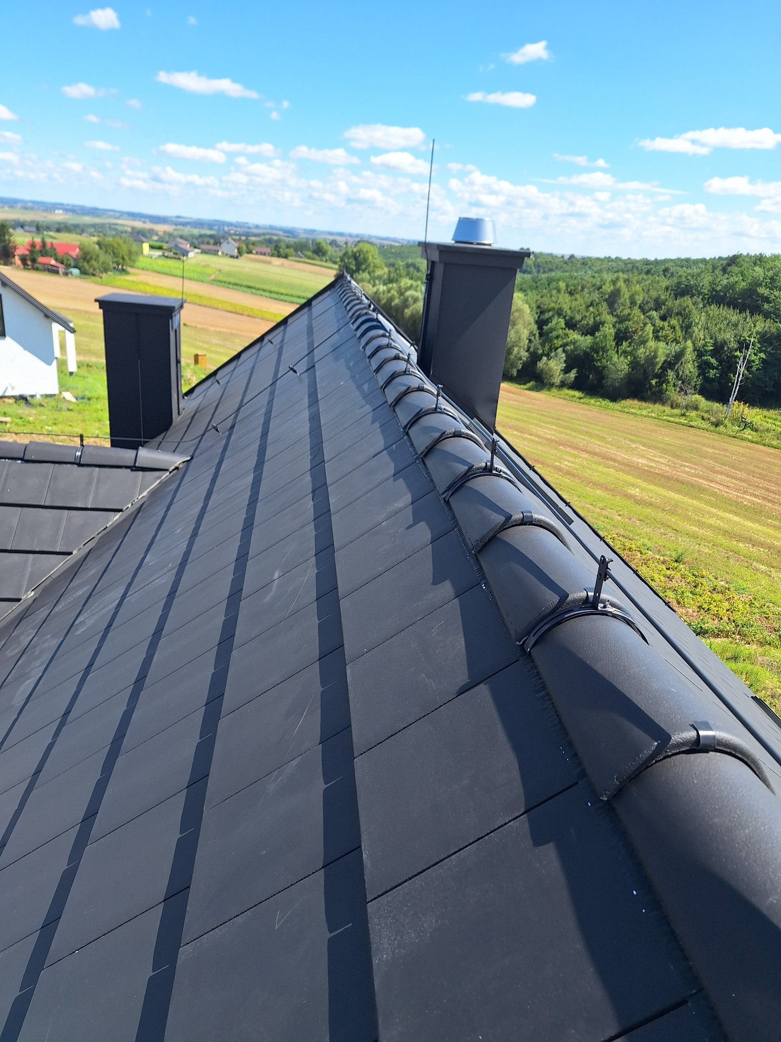 Czarny dach z ceramicznej dachówki karpiówki, z zamontowanymi uchwytami do odgromu, na tle zielonych pól i błękitnego nieba. Widok z góry.