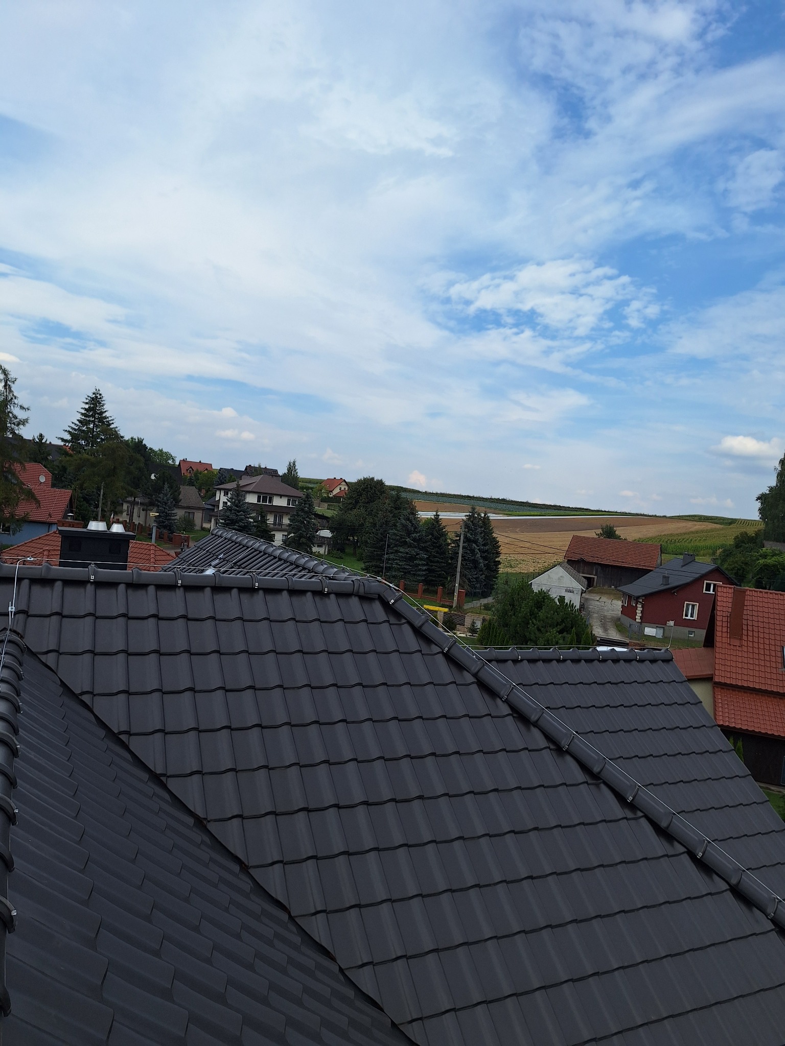 Ciemna dachówka ceramiczna na dachu domu, widok z góry na tle zabudowań i błękitnego nieba z chmurami. Układana w jodełkę, z elementami instalacji odgromowej.