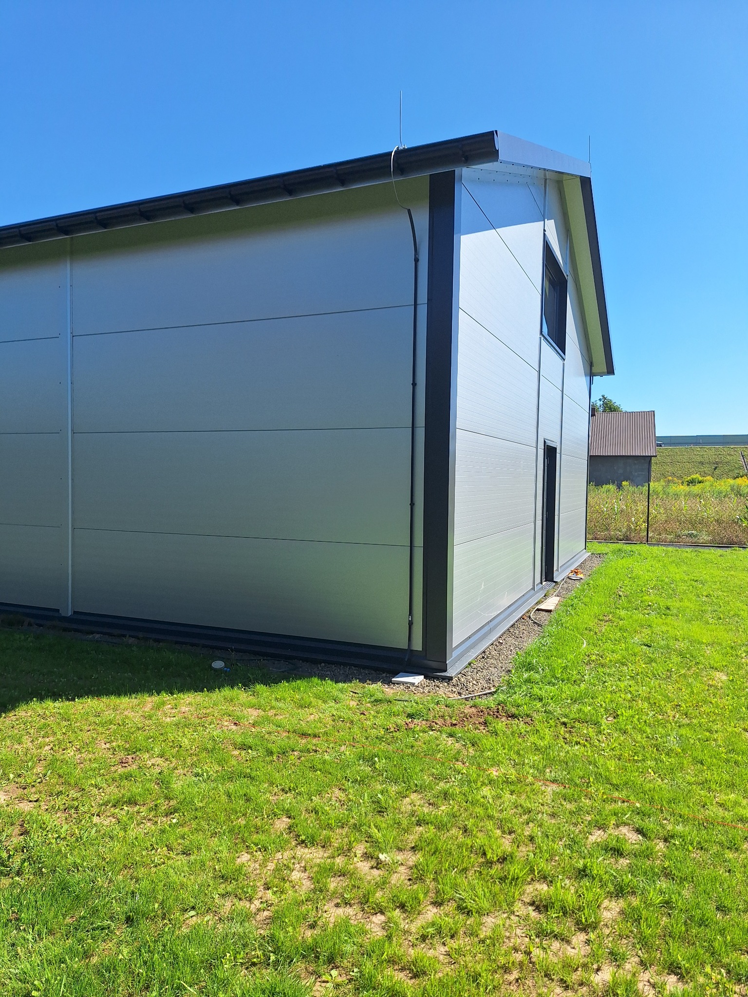 Gotowy budynek modułowy z szarej blachy trapezowej, z czarnymi obramowaniami okien i dachu, na zielonej trawie w słoneczny dzień. Widok z boku, lekko z dołu.