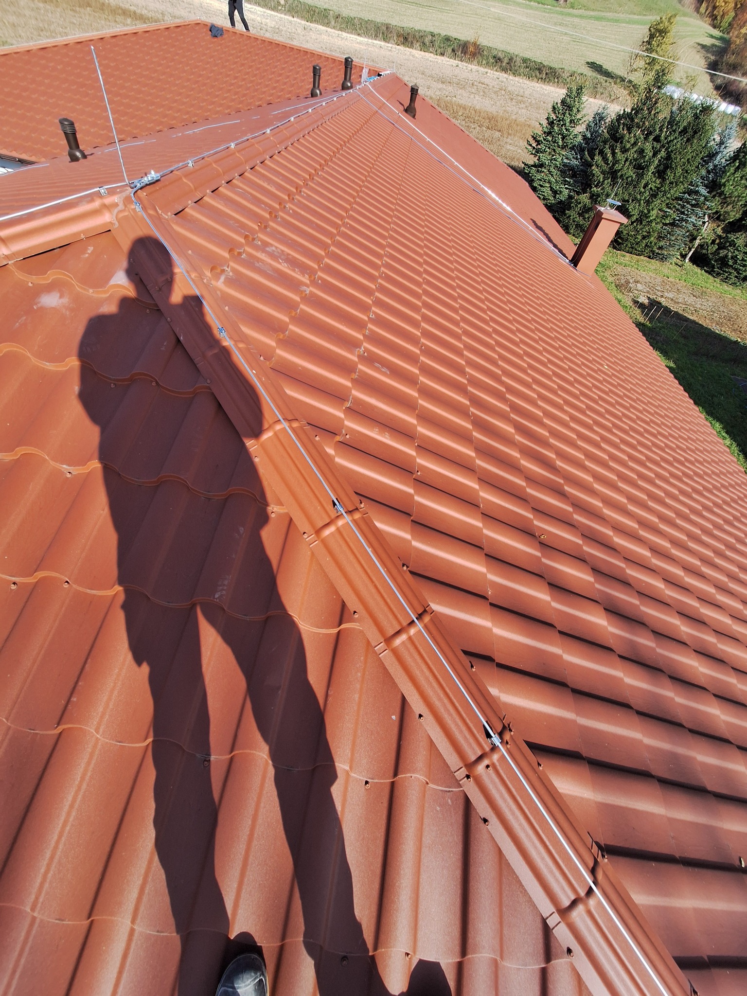 Czerwona dachówka z zamontowaną instalacją odgromową, widok z góry z cieniem osoby, w tle zielony krajobraz.