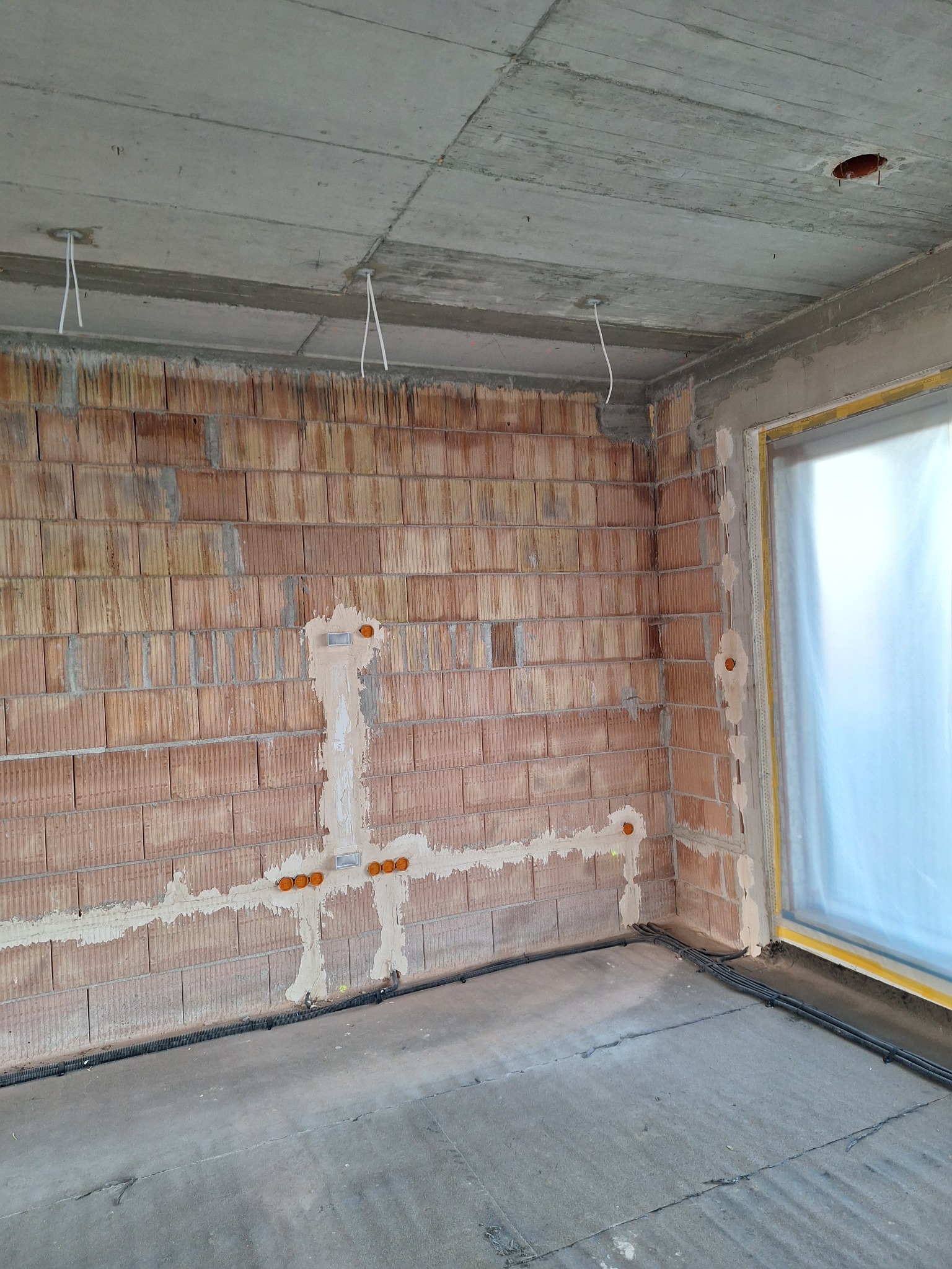 Surowy stan: instalacja elektryczna w ścianie z cegły dziurawki, widoczne puszki i przewody, betonowy sufit z wystającymi kablami, okno zabezpieczone folią.