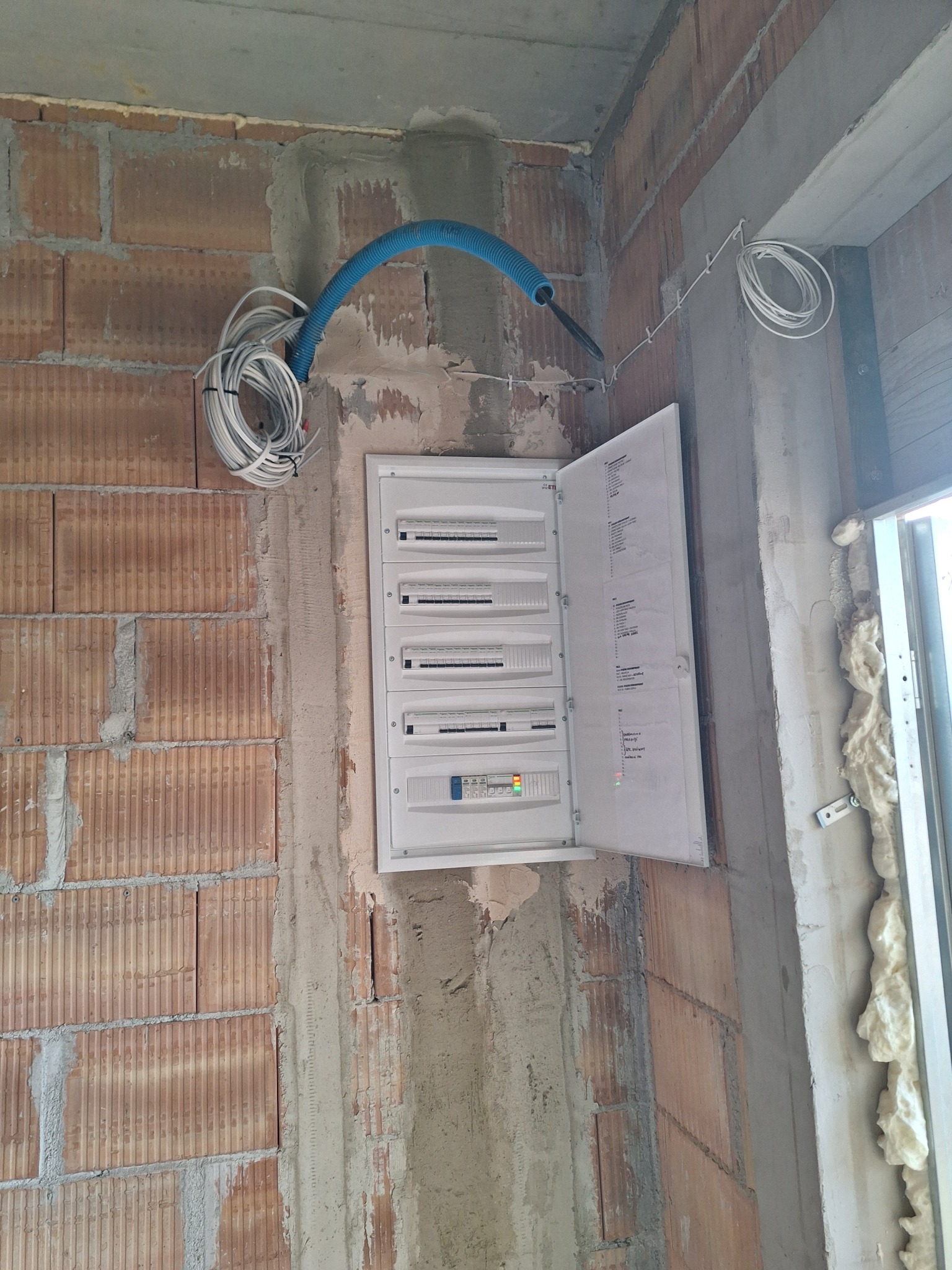 Otwarta skrzynka elektryczna z przewodami na surowej ścianie z cegły. Widoczne elementy instalacji i okno z pianką montażową.