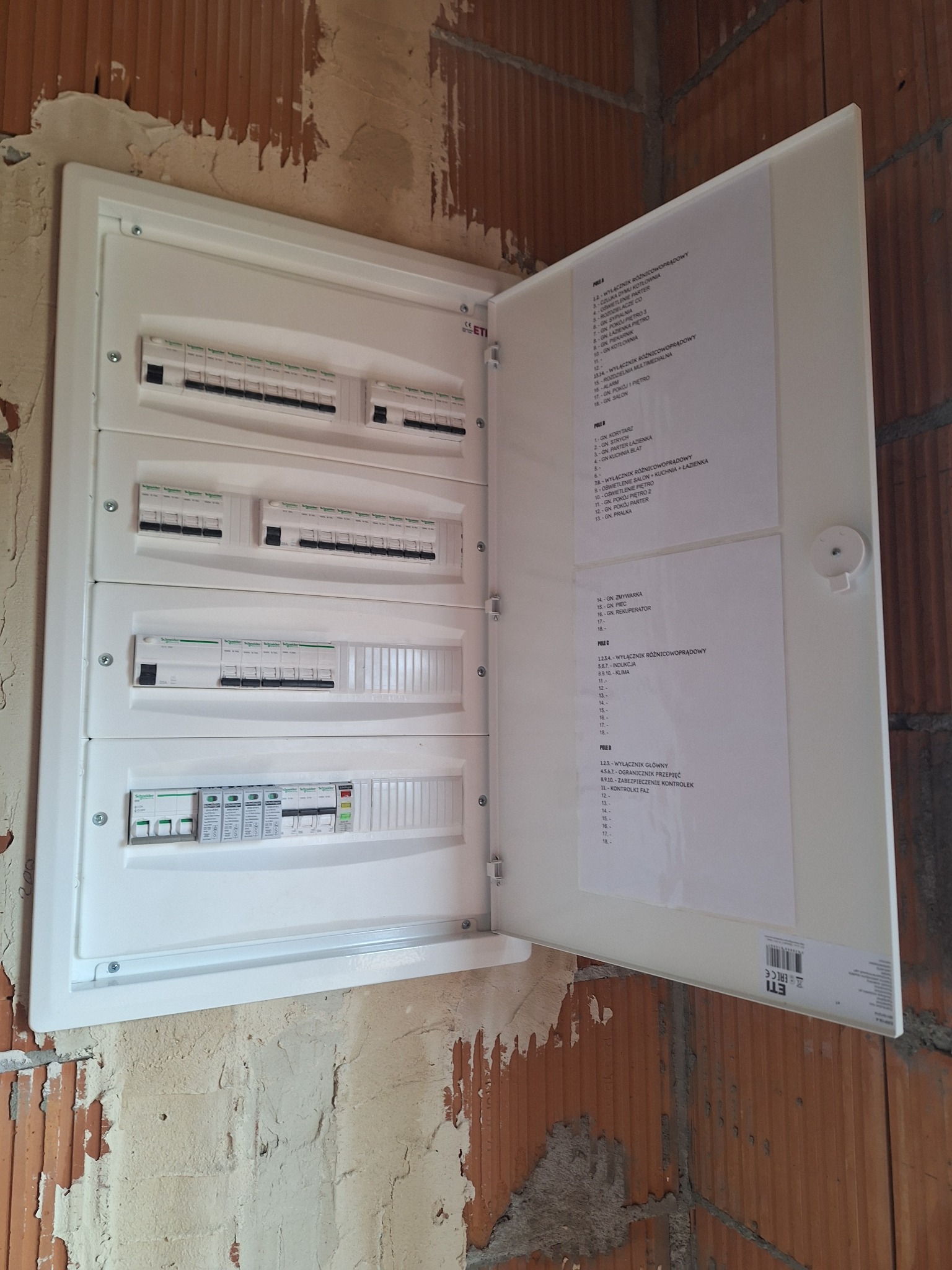 Otwarta, biała skrzynka rozdzielcza z widocznymi bezpiecznikami i opisami, zamontowana na surowej ścianie z czerwonej cegły. Widoczna lista obwodów.