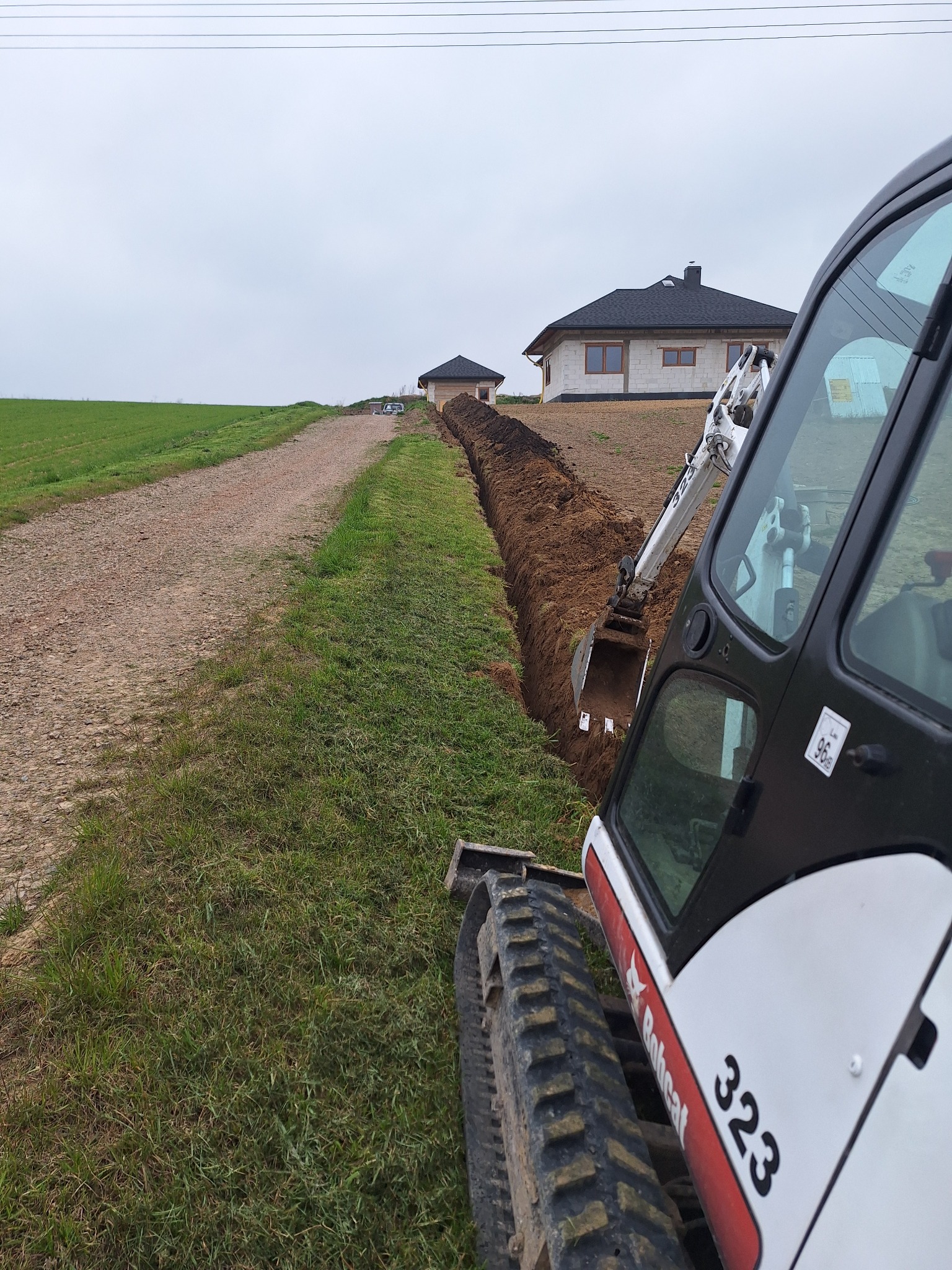 Mini koparka Bobcat 323 wykonuje wykop liniowy na trawiastym terenie, przygotowując podłoże pod instalacje w pobliżu nowo wybudowanego domu z białego pustaka.