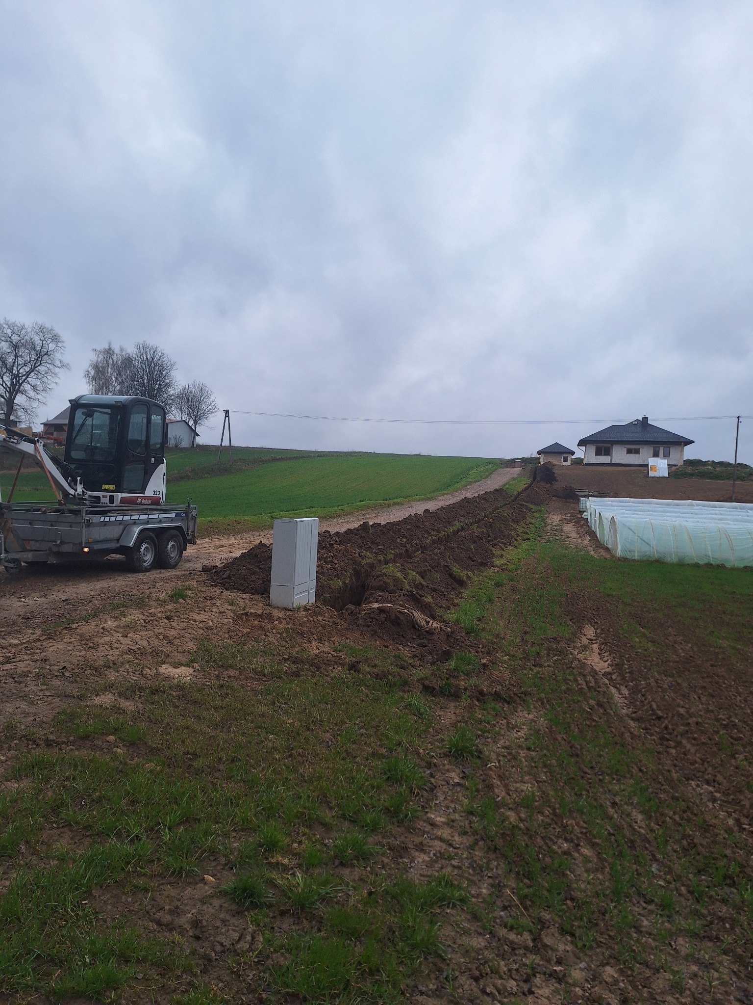 Wykopany rów w ziemi pod instalacje, z widocznym minikoparką na przyczepie i szarą szafką elektryczną. W tle dom i szklarnie.