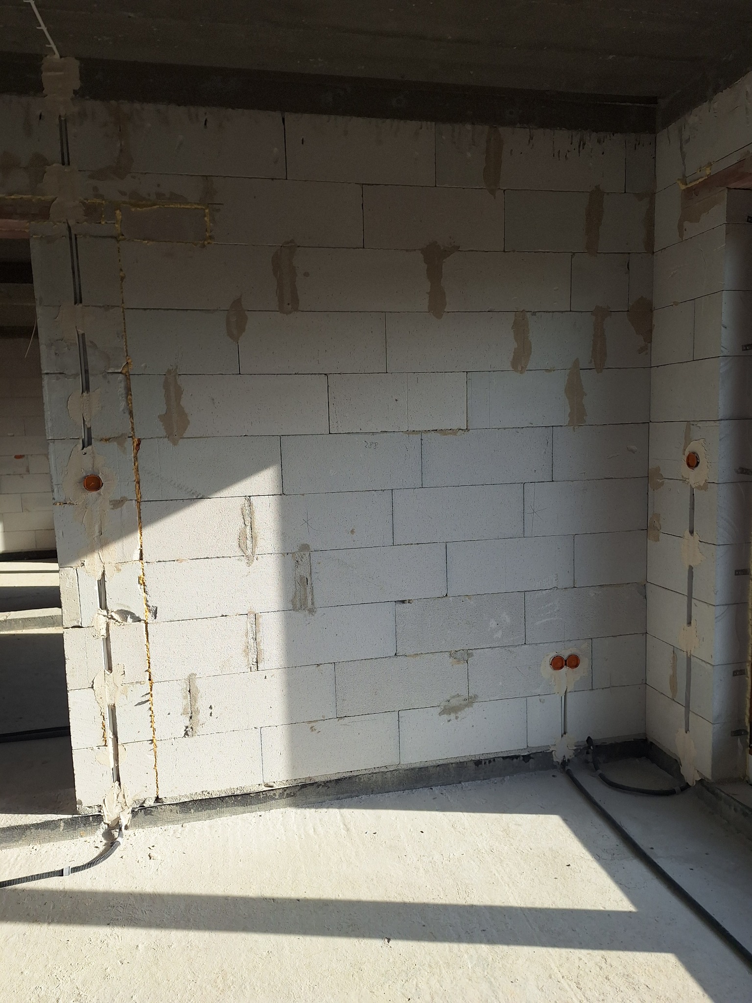 Surowy mur z betonu komórkowego z wyżłobieniami pod instalacje elektryczne i widocznymi puszkami. Kable prowadzone po podłodze. Stan surowy budynku.