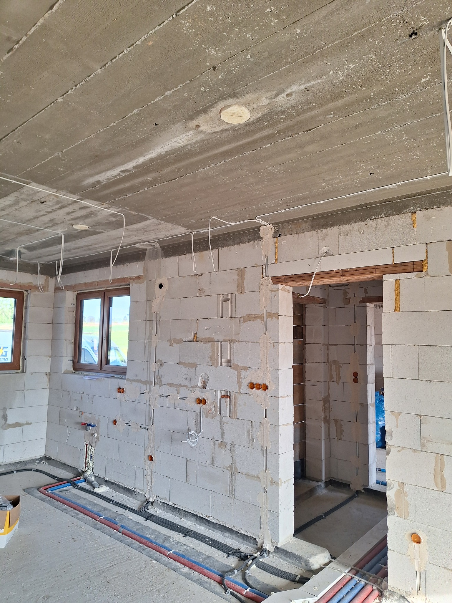 Surowy stan wnętrza domu w trakcie budowy: instalacje elektryczne w ścianach z betonu komórkowego, rury w podłodze, widok na okna i surowy betonowy strop.