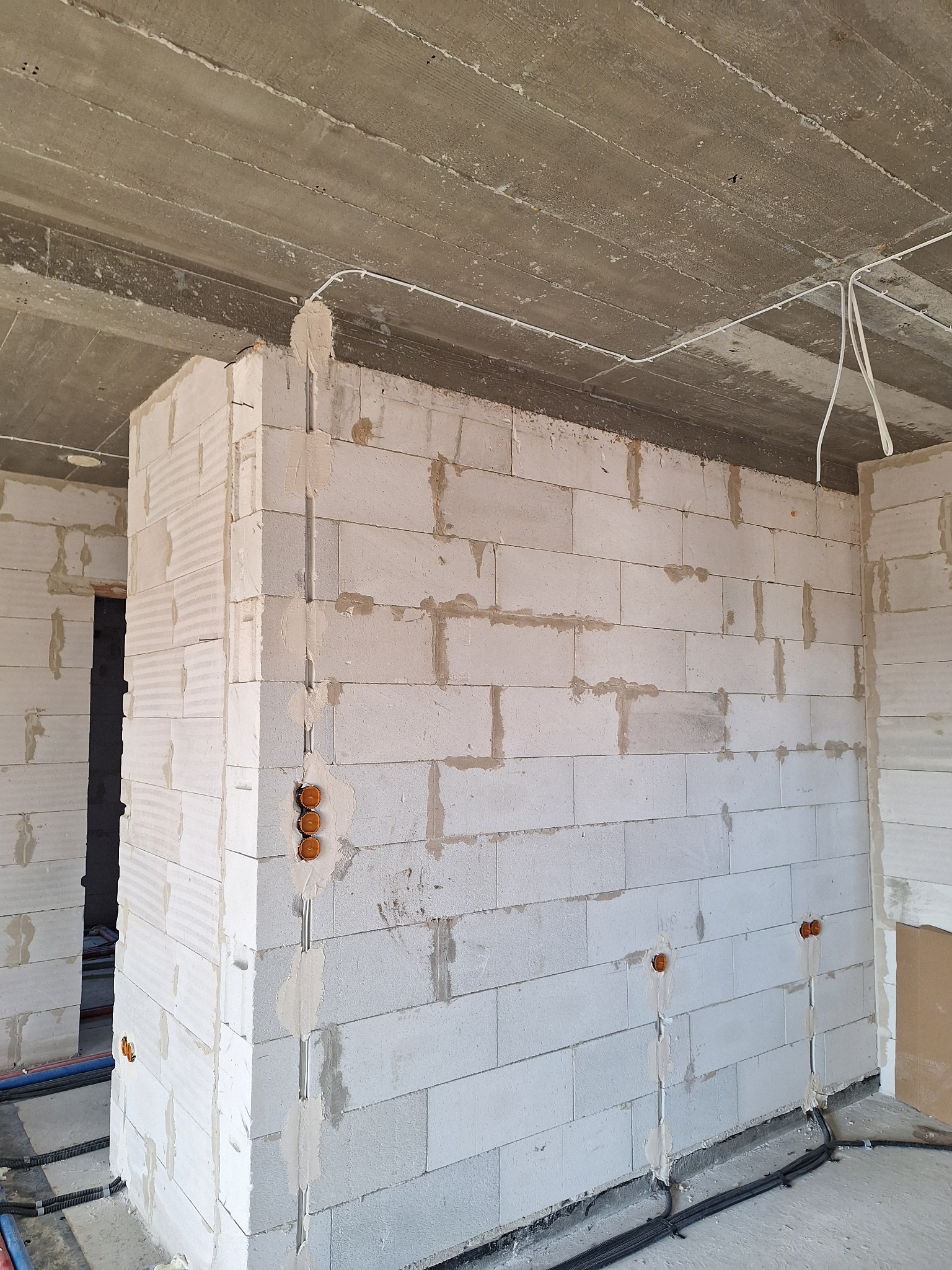 Surowy mur z betonu komórkowego z bruzdami i puszkami pod instalację elektryczną. Widoczne kable i peszle. Szary betonowy strop.