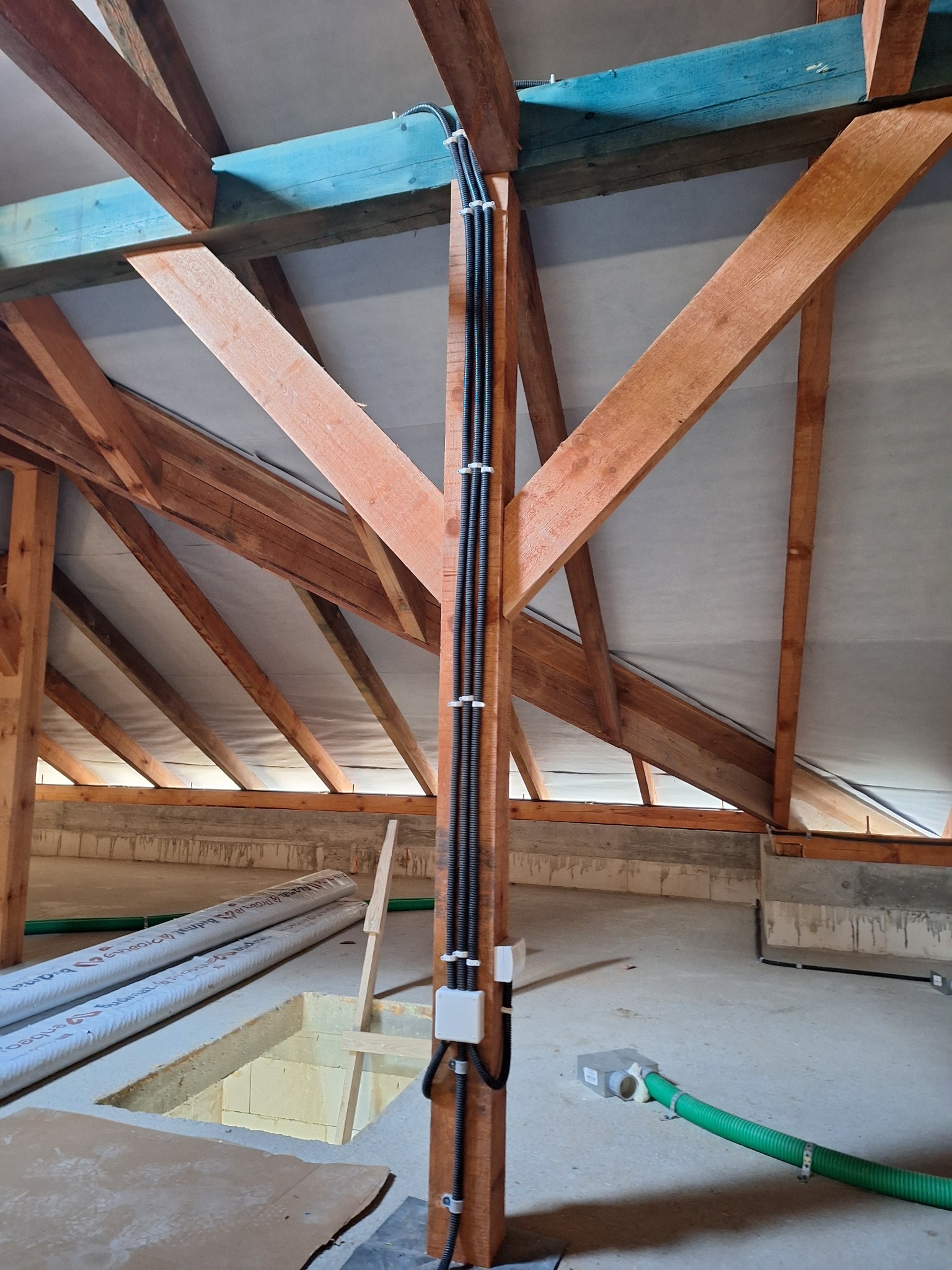 Wewnętrzna instalacja elektryczna w budynku w trakcie budowy. Kable w peszlach przymocowane do drewnianej konstrukcji dachu. Widoczne puszki elektryczne i elementy wentylacji.