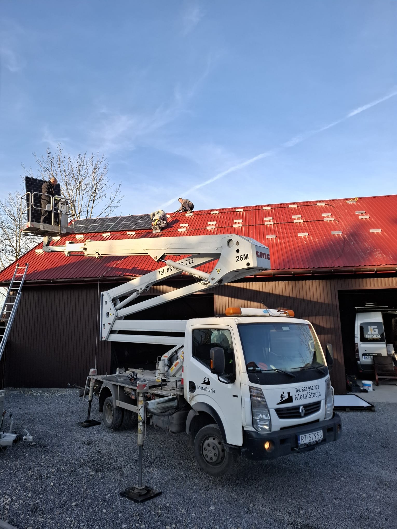 Montaż paneli słonecznych na czerwonym dachu budynku gospodarczego z użyciem podnośnika koszowego. Widoczny samochód dostawczy firmy MetalStacja.