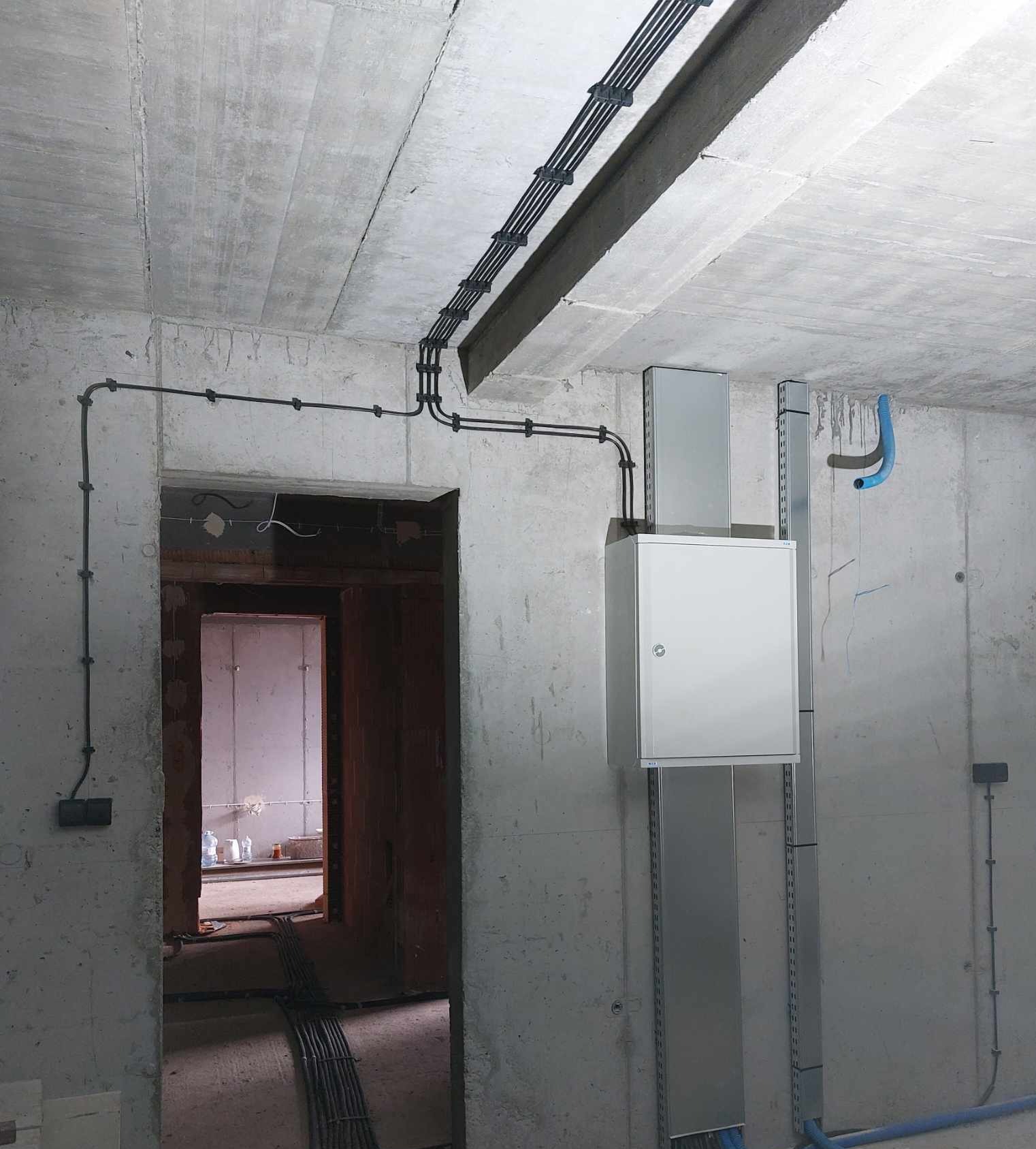 Surowy beton i instalacje elektryczne: kable w peszlach na suficie, puszka rozdzielcza na ścianie, widok z wnętrza budynku w stanie deweloperskim.