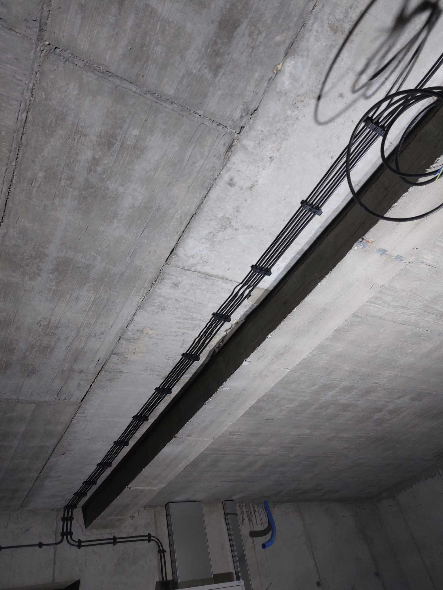 Surowe wnętrze z betonowym stropem, gdzie widoczne są czarne kable elektryczne, równolegle przymocowane do sufitu za pomocą uchwytów. Instalacja w trakcie realizacji.