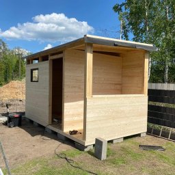 Tarasywwl - Budowa drewnianej konstrukcji z widocznym fragmentem dachu pokrytego papą, narzędzia leżące na trawie i drabina oparta o ścianę, w tle zielone drzewa i błękitne niebo z chmurami.