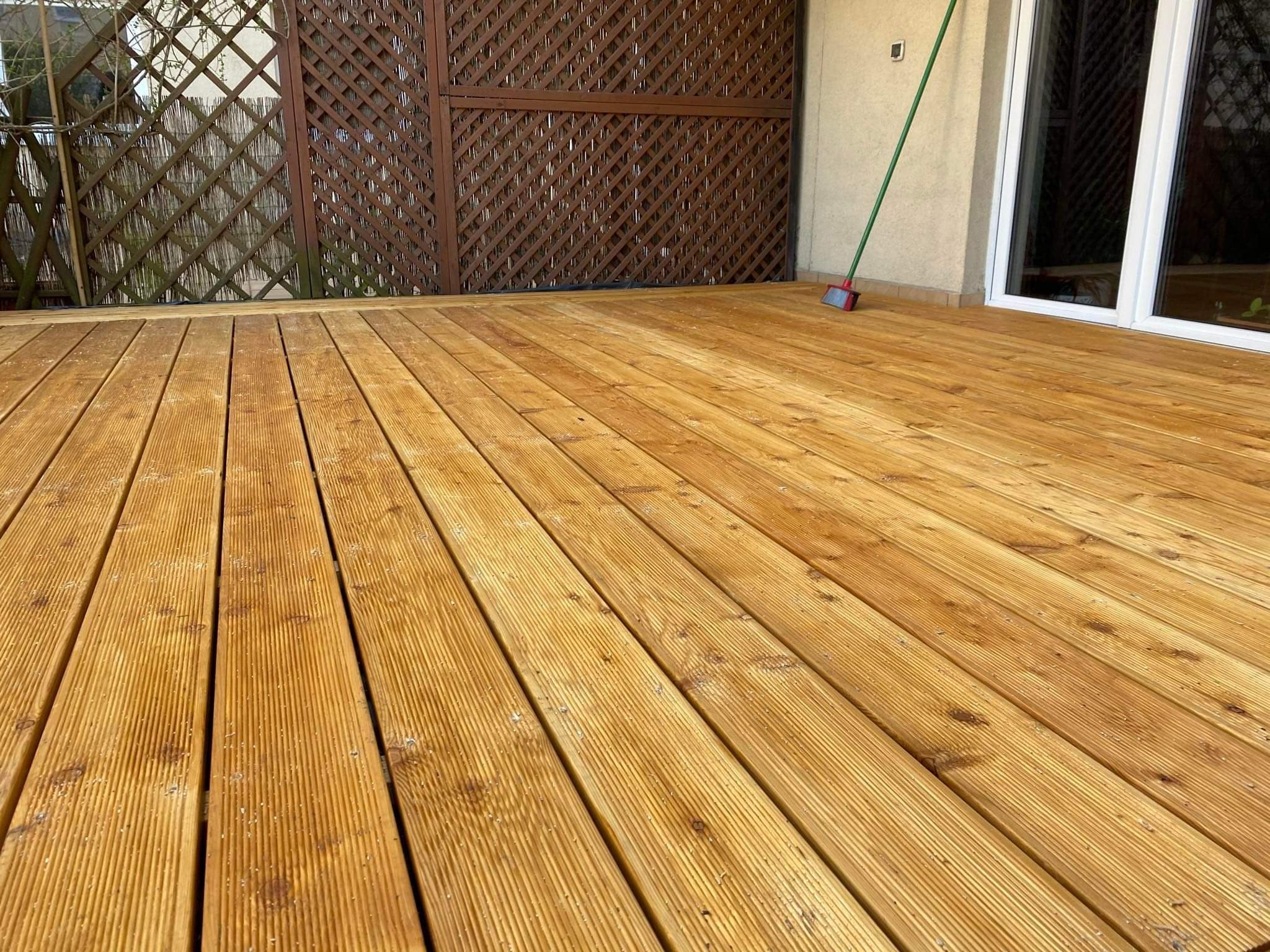 Świeżo wyczyszczony taras drewniany z widoczną fakturą desek, z brązową ażurową zabudową i mopem opartym o ścianę budynku.