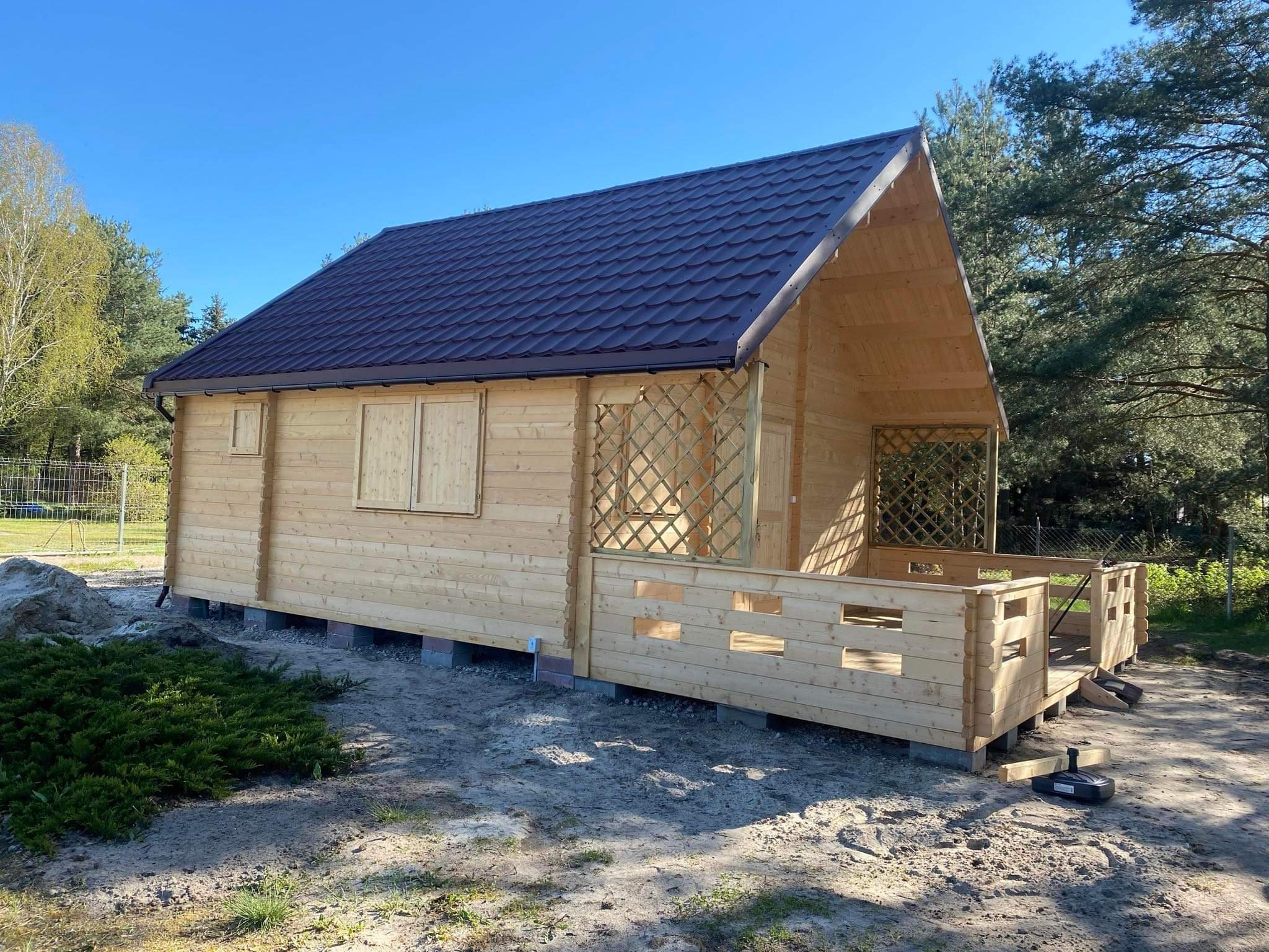 Surowa drewniana konstrukcja altany z brązowym dachem, widoczna w trakcie montażu na betonowych blokach, otoczona piaskiem i zielenią.