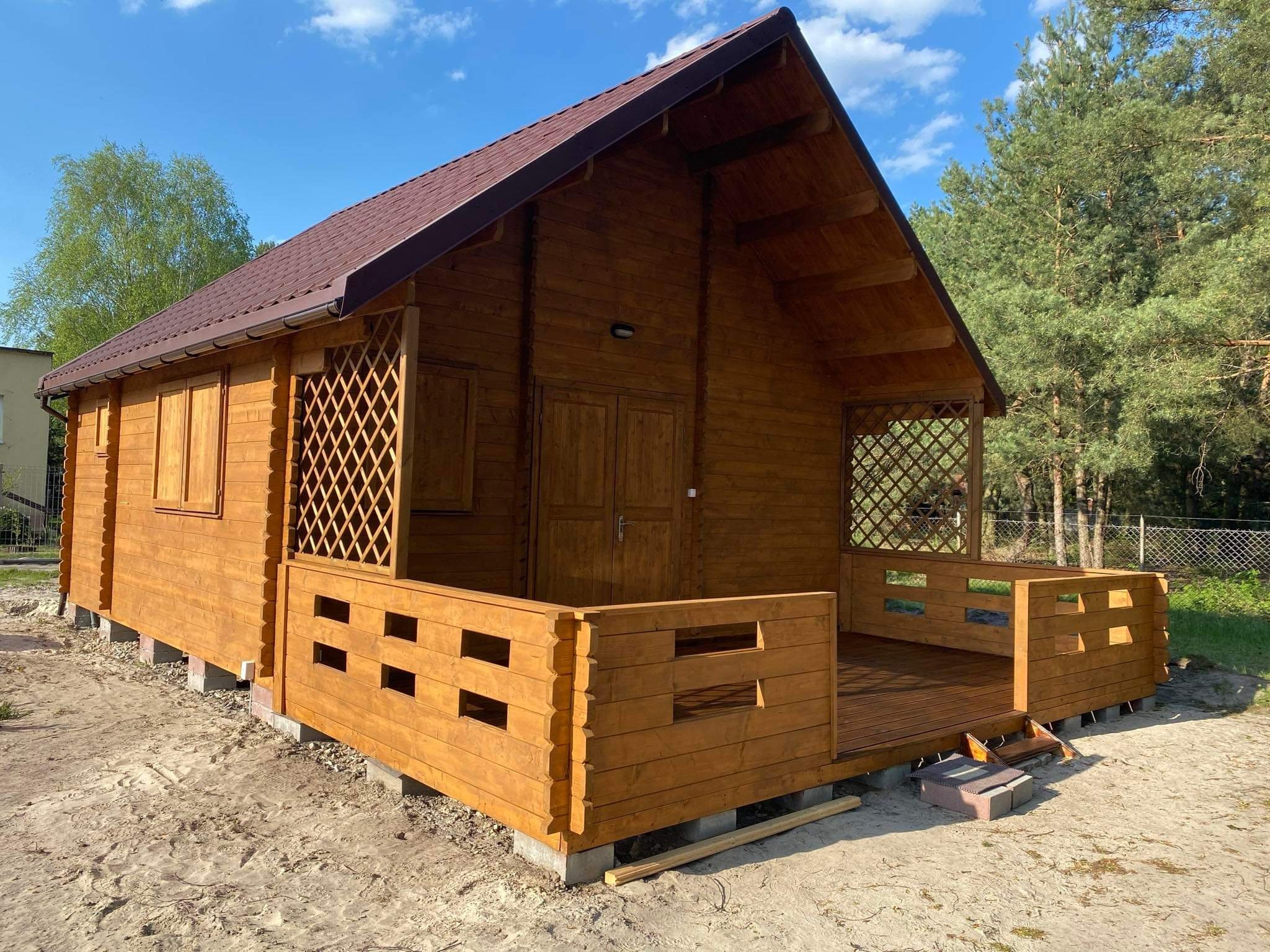 Nowa, drewniana, brązowa altana z kratownicą i małym tarasem, osadzona na betonowych bloczkach, na piaszczystym podłożu, z widocznymi drzewami w tle.