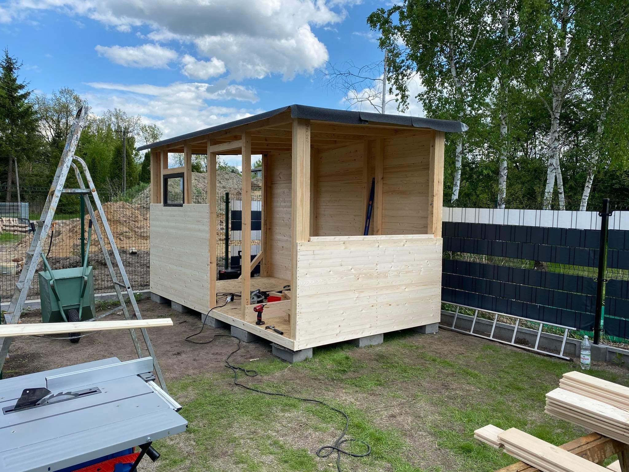 Konstrukcja drewnianej altany w trakcie budowy, widoczne elementy szkieletu, deski obiciowe, narzędzia, drabina i piła stołowa na trawniku.