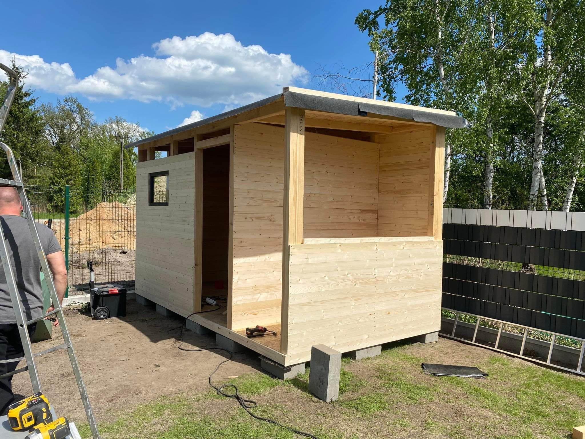 Budowa drewnianej konstrukcji z widocznym fragmentem dachu pokrytego papą, narzędzia leżące na trawie i drabina oparta o ścianę, w tle zielone drzewa i błękitne niebo z chmurami.