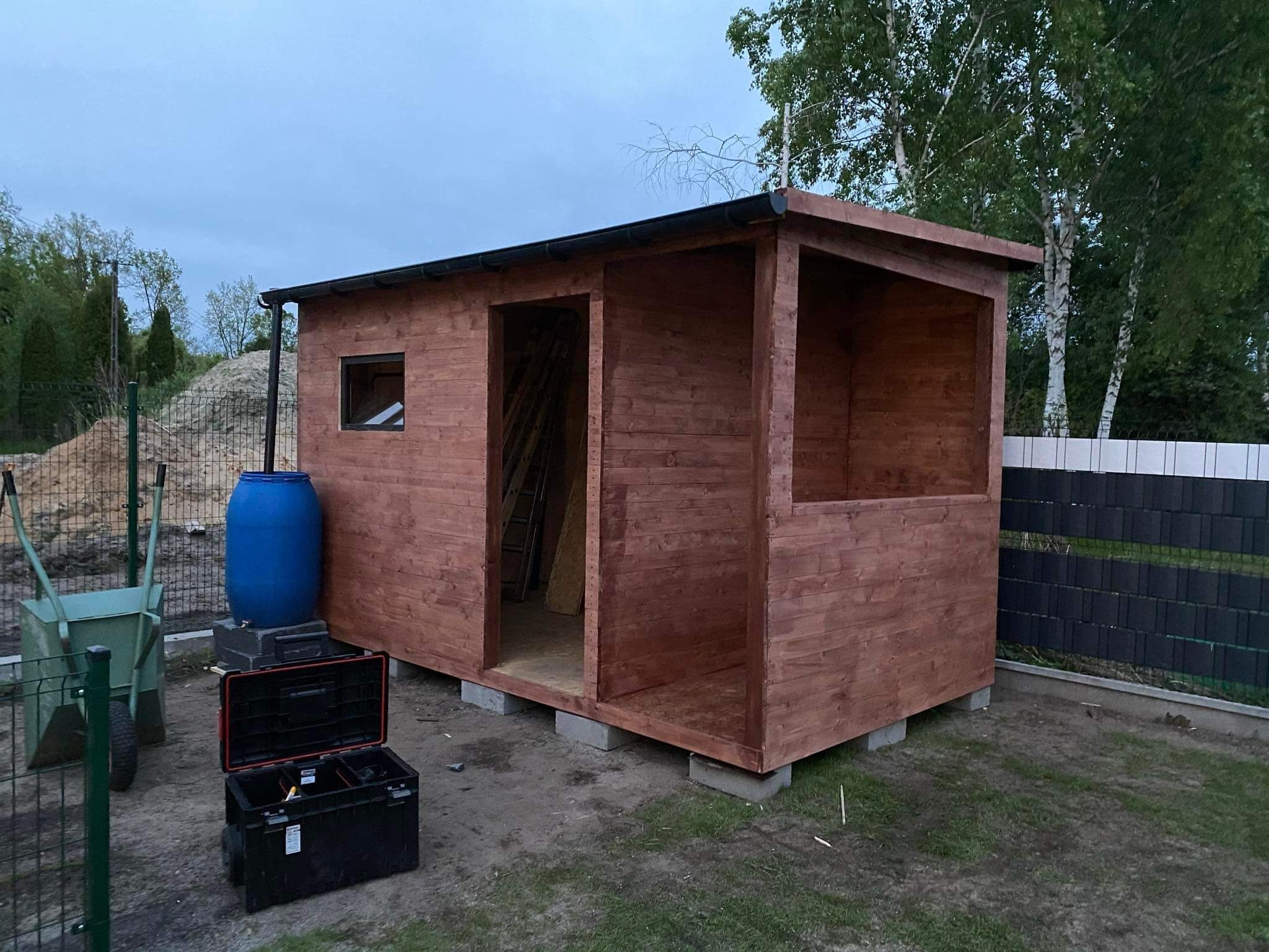 Drewniana konstrukcja z oknem i otwartą przestrzenią, prawdopodobnie w trakcie budowy, stoi na betonowych bloczkach, obok widoczny wózek, beczka i skrzynka narzędziowa.