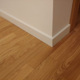 FloorWood Przemysław Wójcik - Jasne panele podłogowe ułożone przy białej ścianie i listwie przypodłogowej, widoczny fragment drewnianej ościeżnicy drzwiowej.