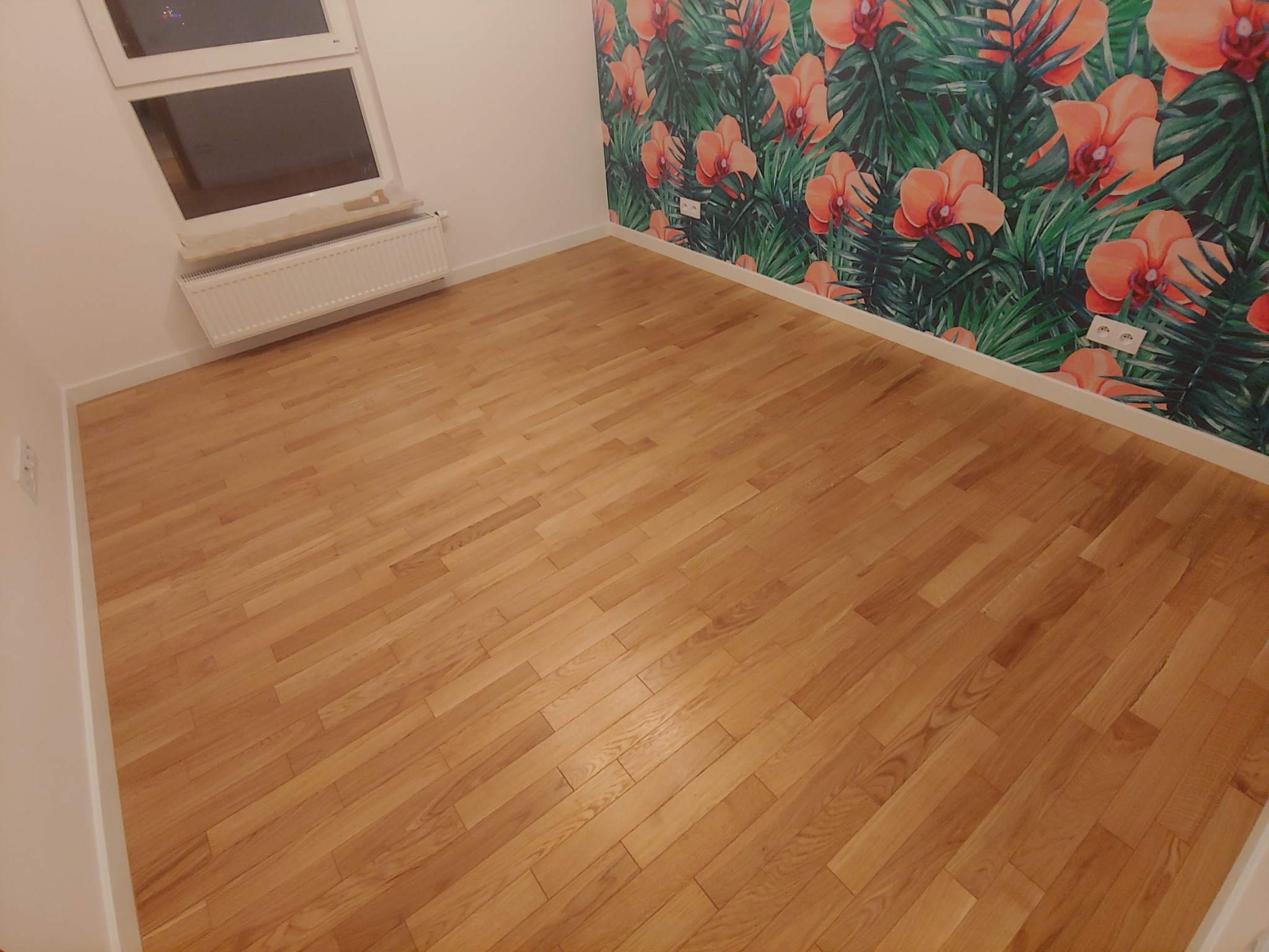 Jasny parkiet ułożony w nowym pokoju z kwiatową tapetą, widoczne gniazdka elektryczne przy podłodze.