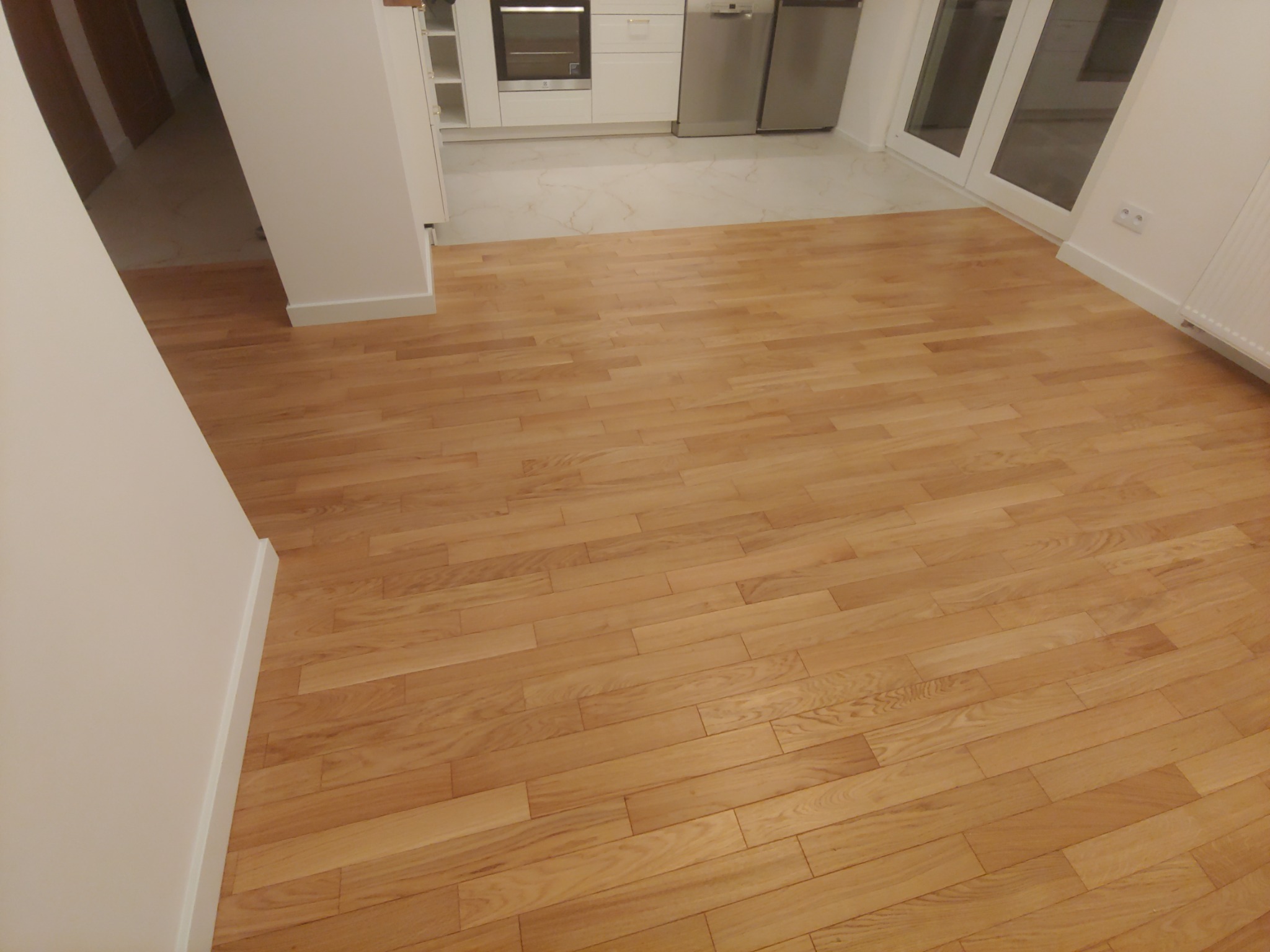 Jasny, drewniany parkiet ułożony w jodełkę francuską w nowocześnie wykończonym mieszkaniu z białą kuchnią i dużymi oknami balkonowymi.