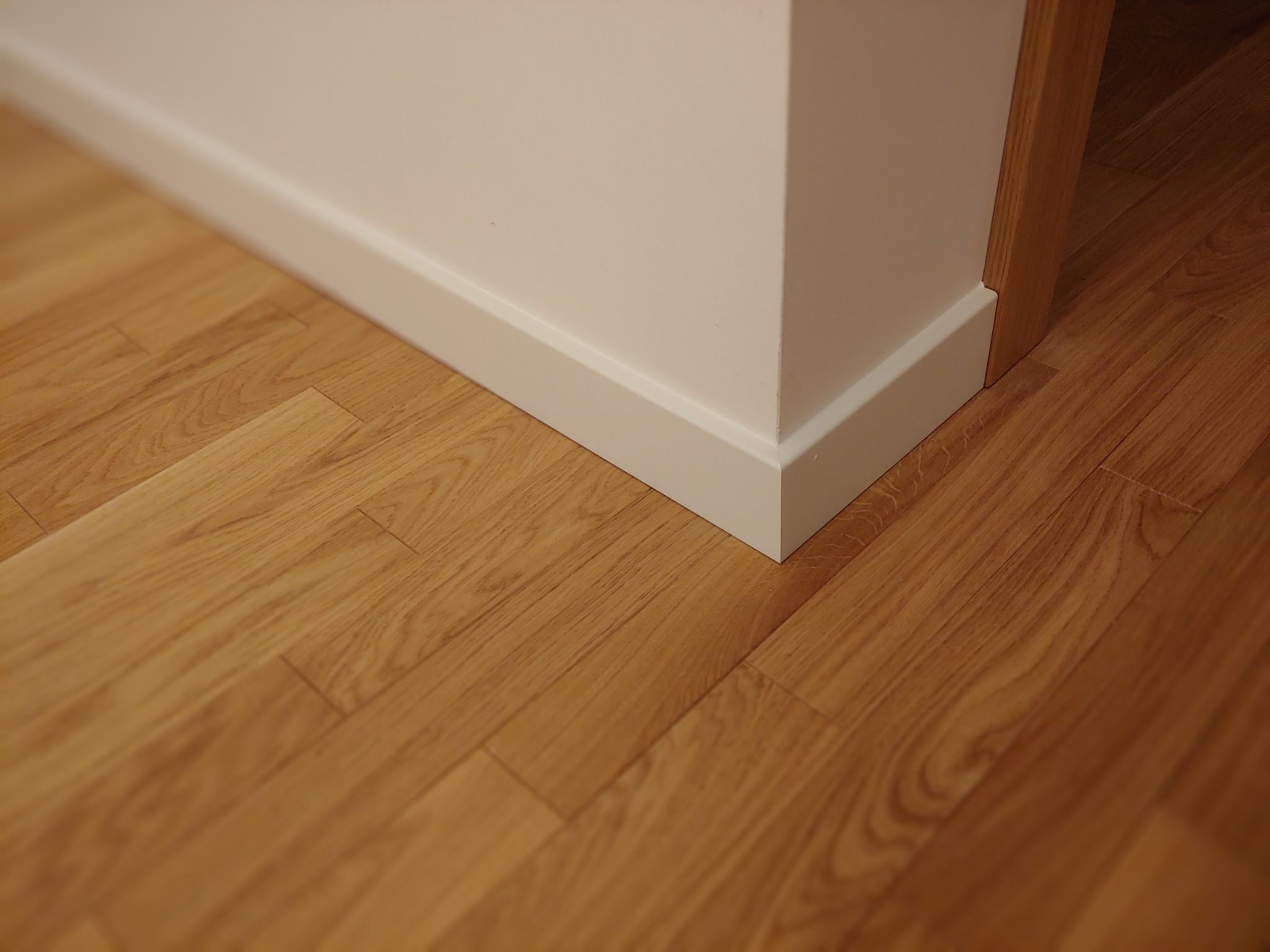 Jasne panele podłogowe ułożone przy białej ścianie i listwie przypodłogowej, widoczny fragment drewnianej ościeżnicy drzwiowej.