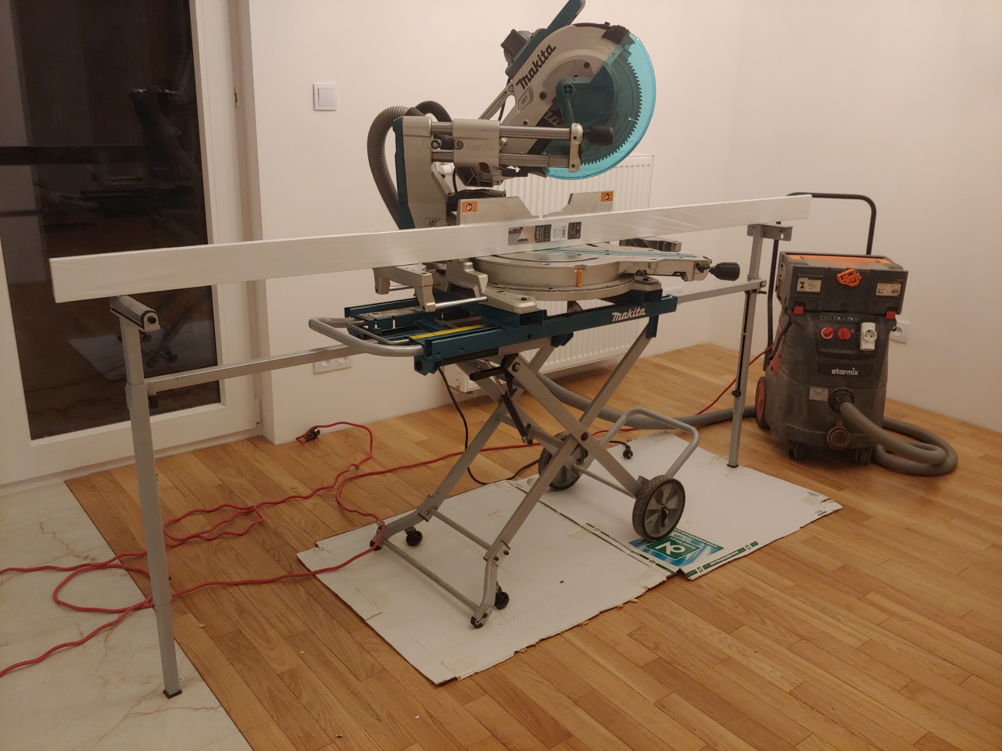 Piła ukosowa Makita na metalowym stojaku, przygotowana do cięcia białej listwy przypodłogowej, obok odkurzacz przemysłowy Starmix, podłoga częściowo zabezpieczona kartonem.