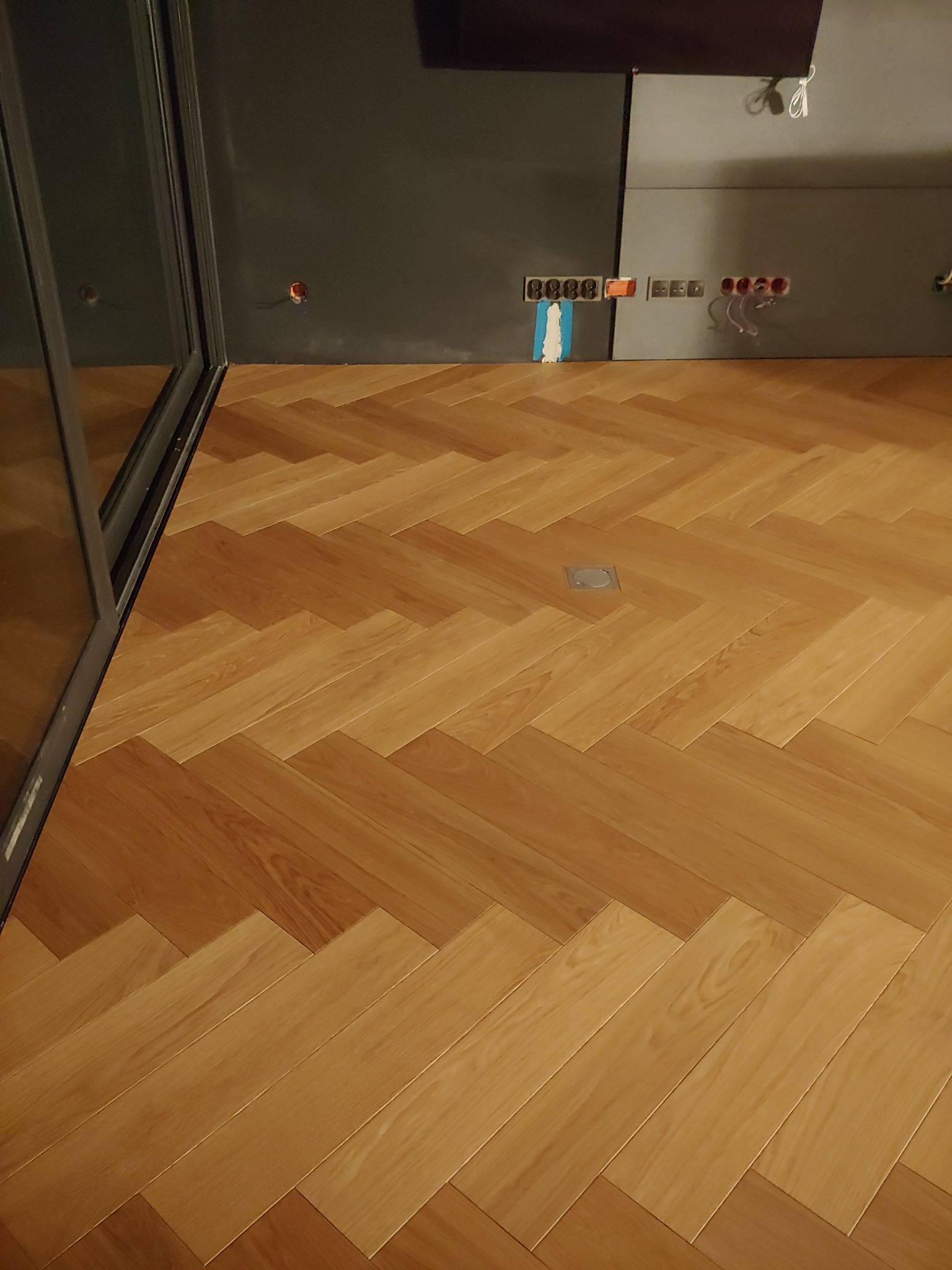 Jasny parkiet ułożony we wzór jodełki francuskiej, widoczny fragment ściany z wyprowadzonymi instalacjami elektrycznymi.